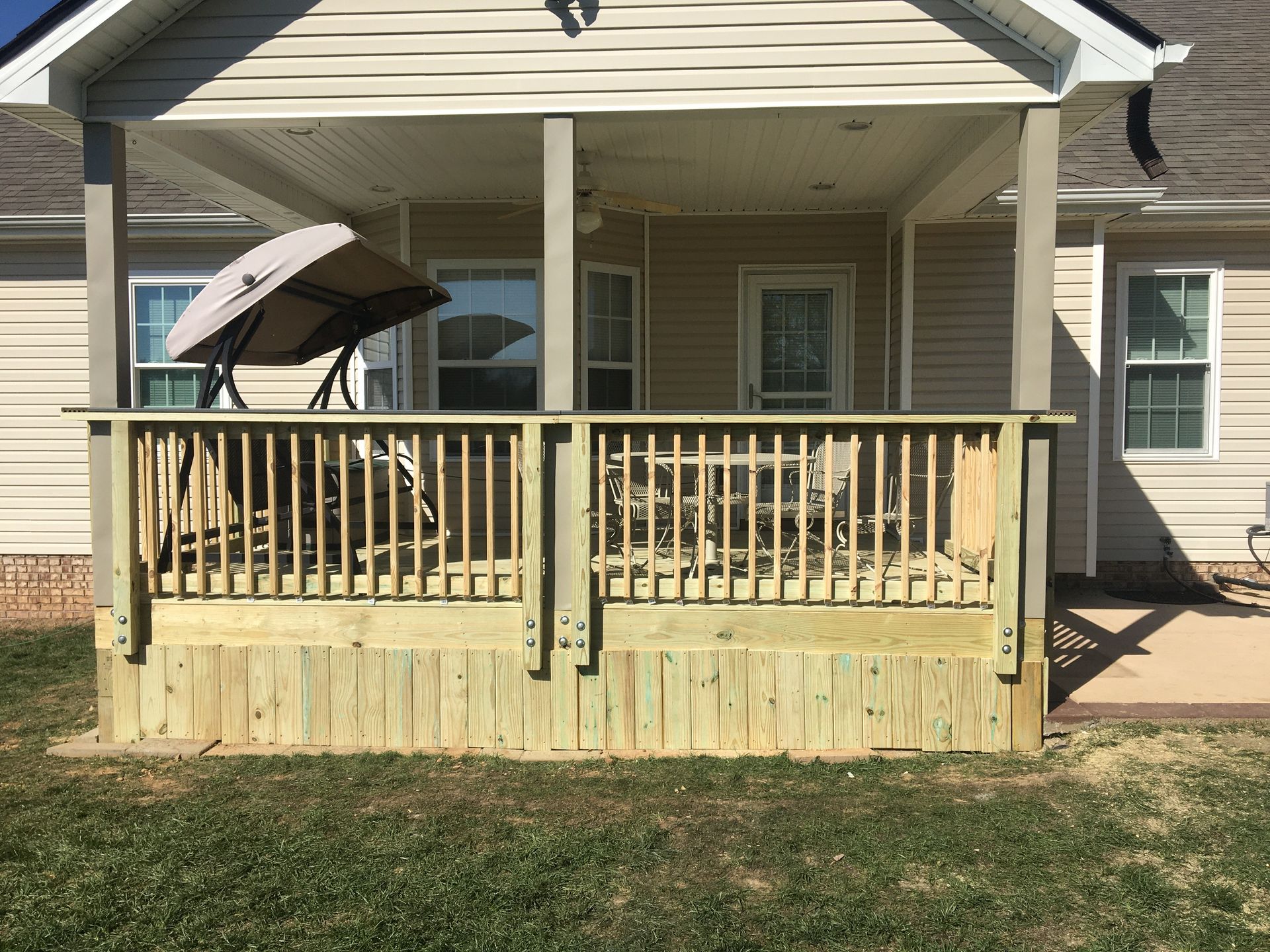 Wooden deck with railing attached to a beige house, with an umbrella, swing, and chairs.
