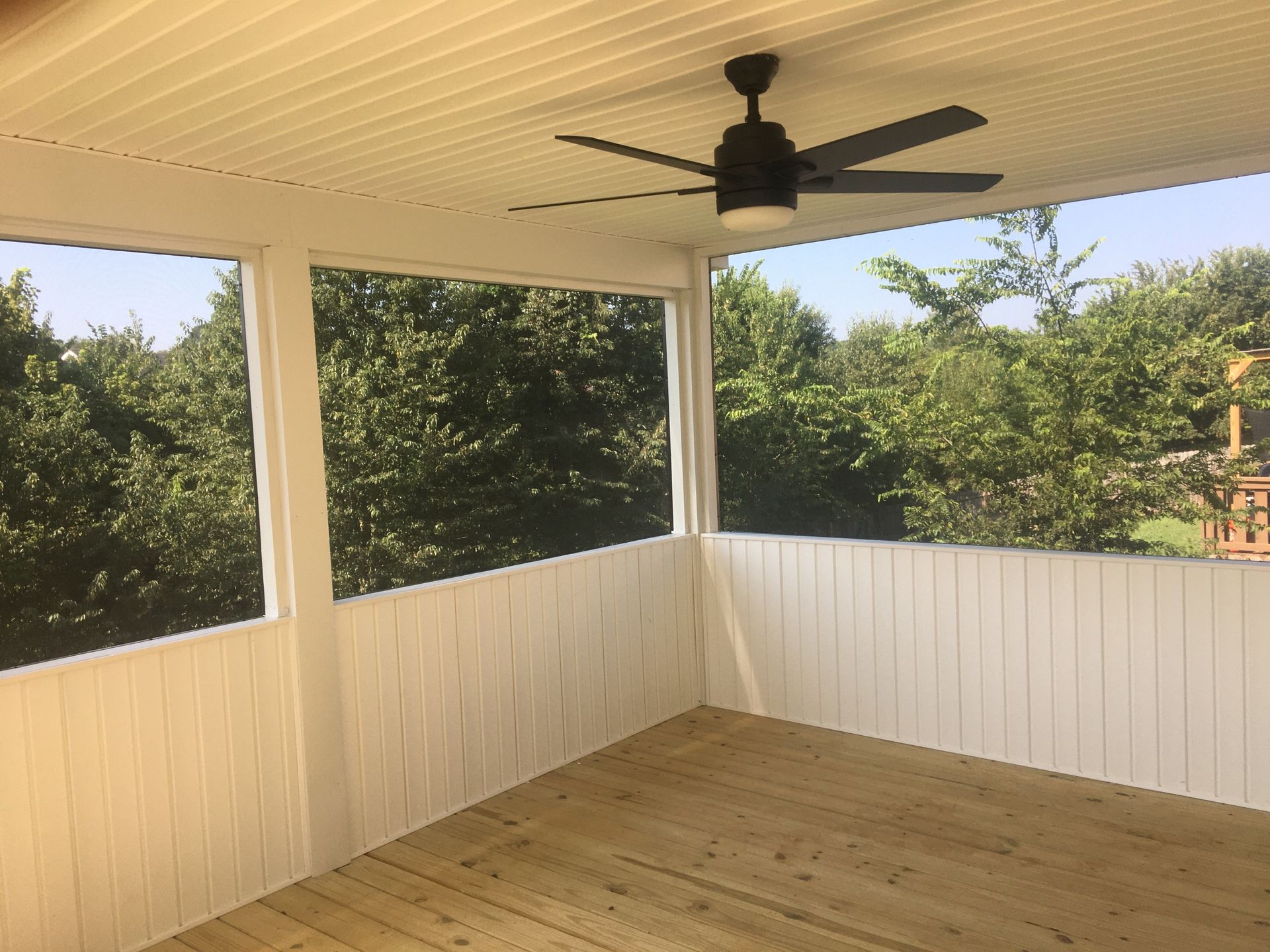 Screened-in porch with white walls and ceiling, brown floor, ceiling fan, and view of trees.