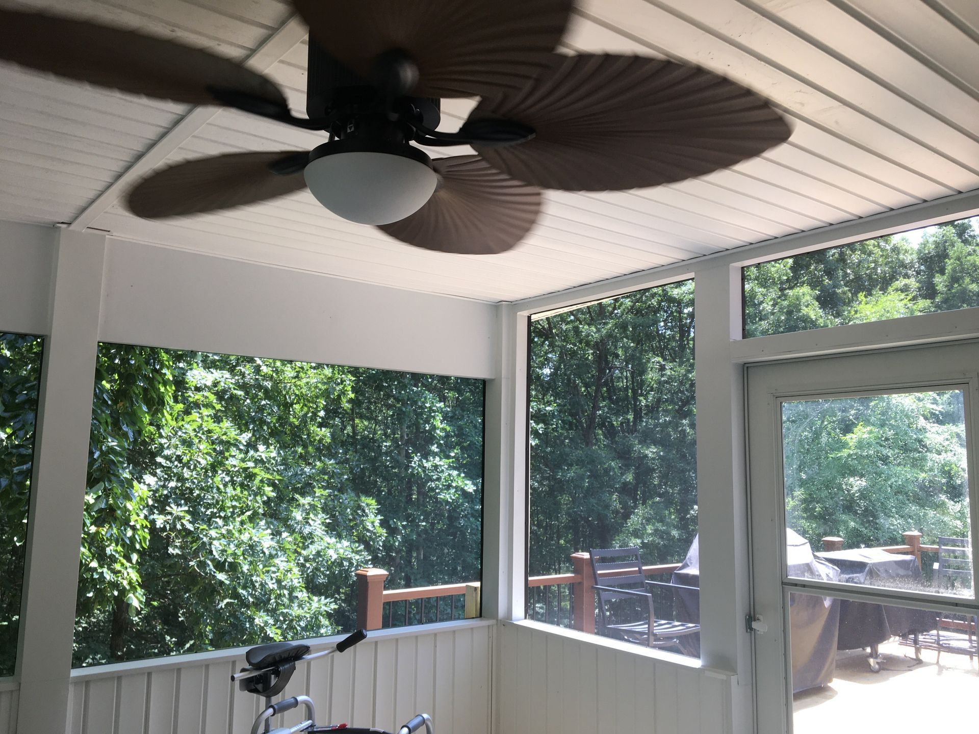 Screened-in porch with ceiling fan, white trim, and views of lush green trees outdoors.