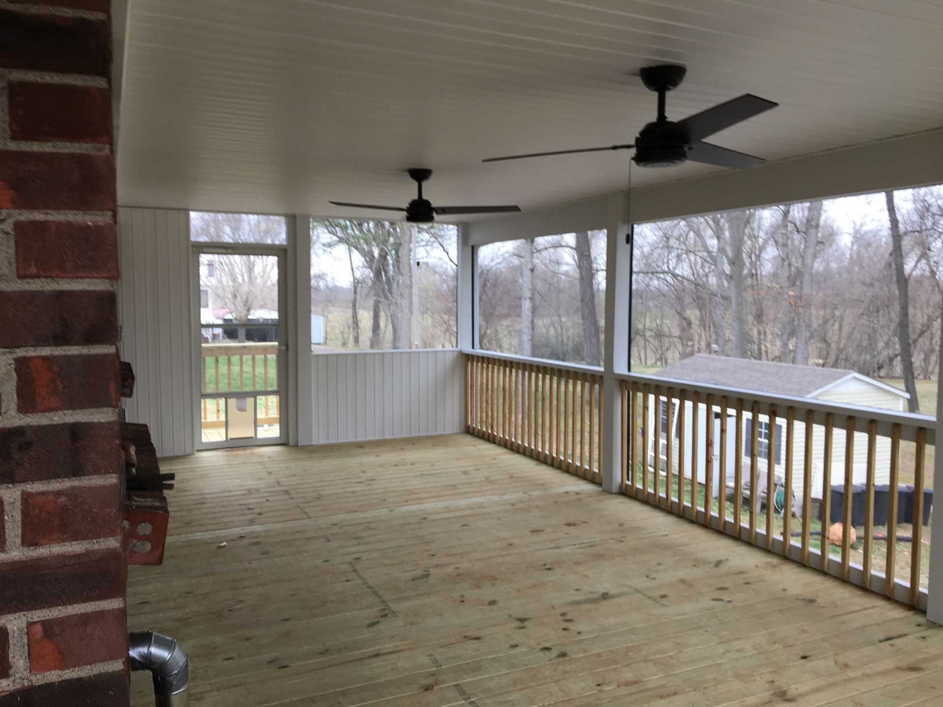 Screened-in porch with wood deck, railing, and ceiling fans. A brick wall is on the left; a view of trees and houses is in the background.