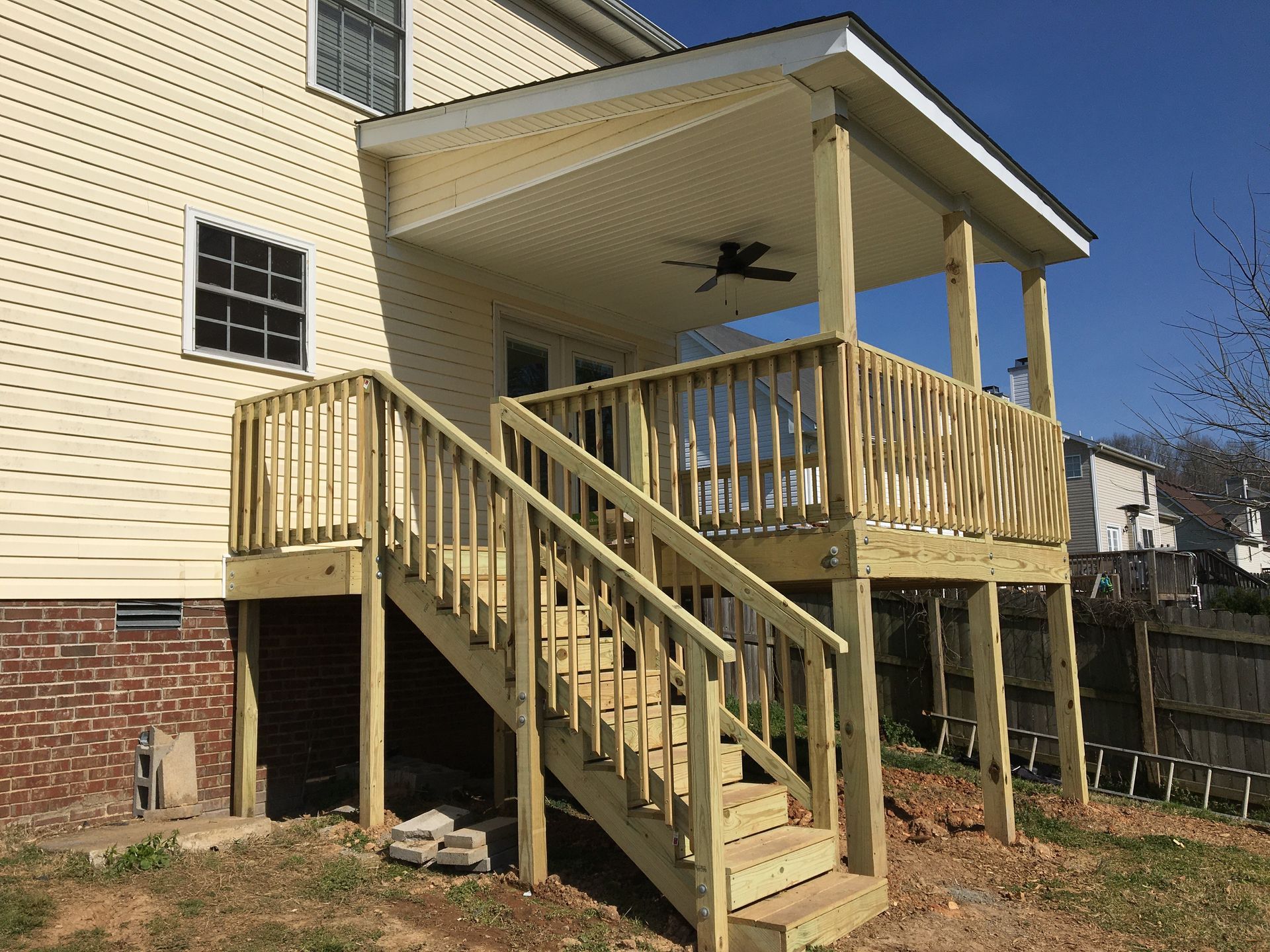 Wooden deck with stairs attached to a two-story house, a covered porch, and a blue sky.