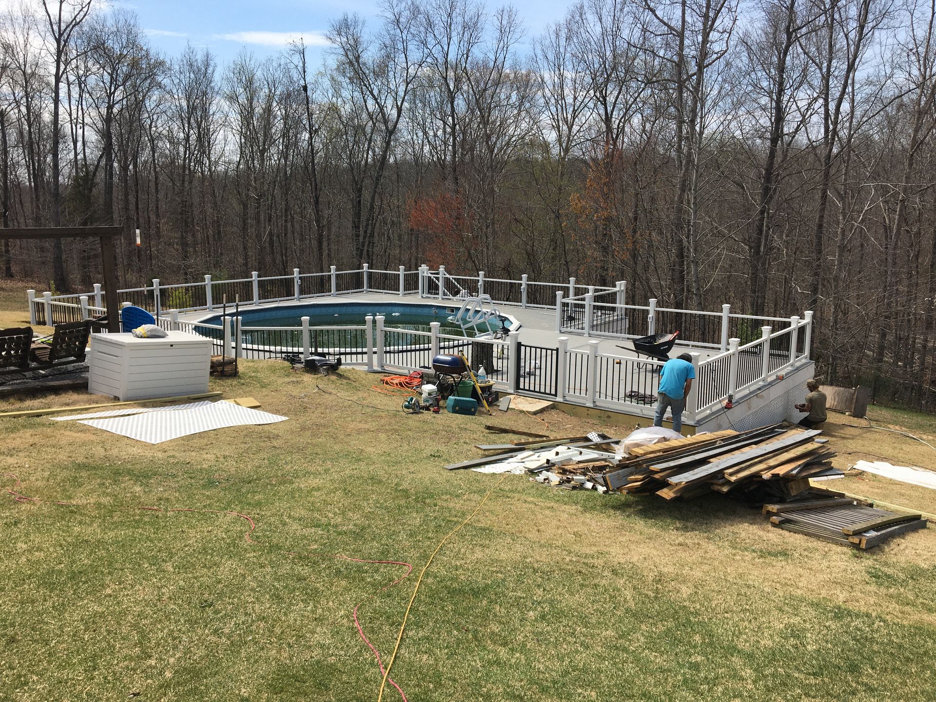 Workers building a deck around an above-ground pool in a grassy backyard, surrounded by trees.