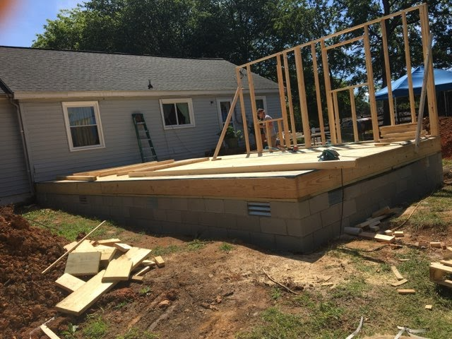 Construction site: deck addition to a light gray house; wood framing and concrete foundation in progress.