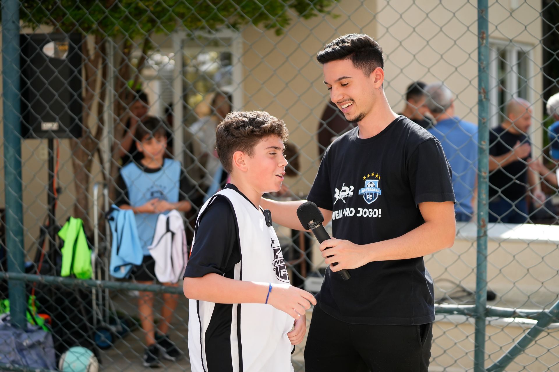 Homem entrevistando um menino com um microfone em um campo de futebol. Ambos estão sorrindo. Outras pessoas e uma cerca são visíveis.
