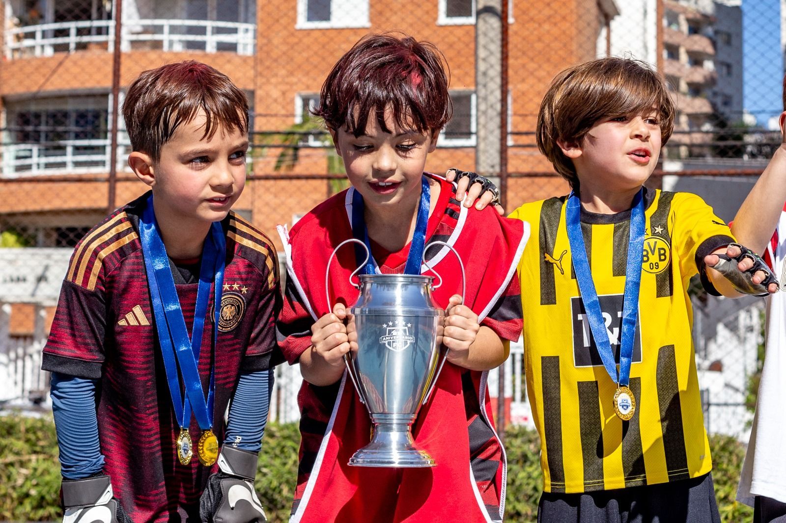 Três jovens jogadores de futebol com medalhas, segurando um troféu, comemorando ao ar livre.