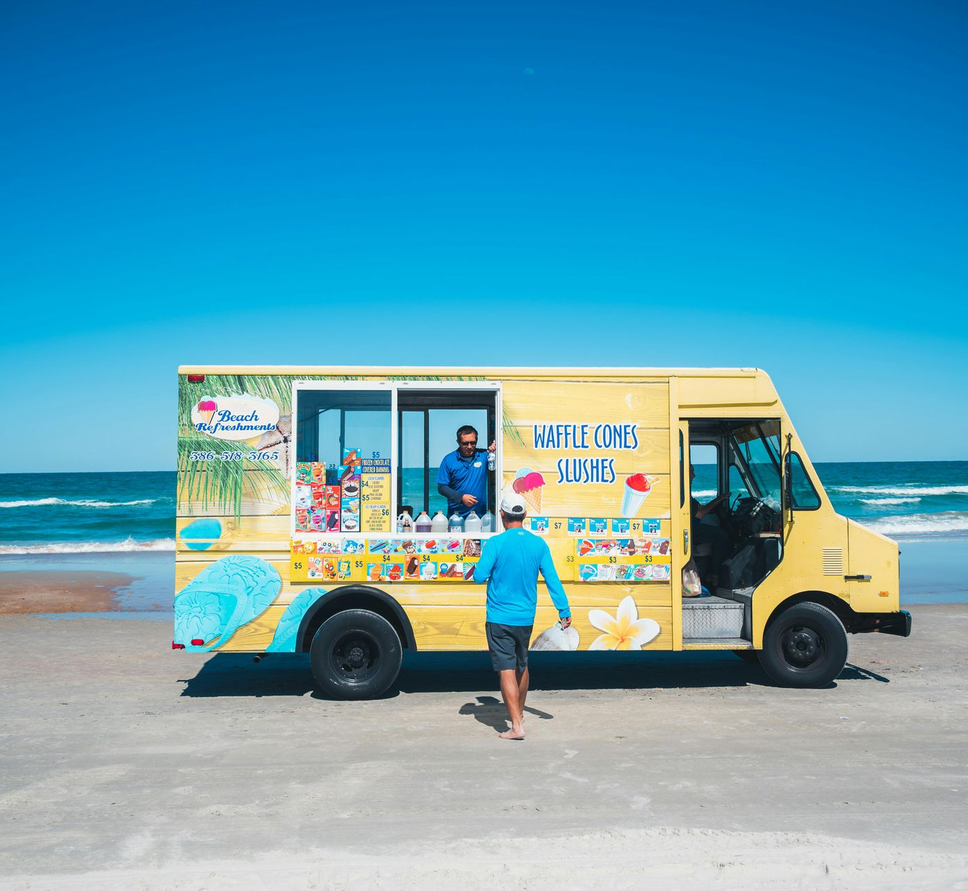 Yellow ice cream truck on a beach, with a person standing at the serving window under a bright blue sky