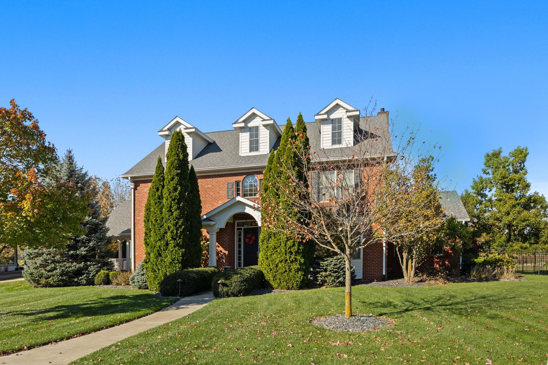 Large brick house with gray roof, tall evergreens, and a green lawn under a clear blue sky