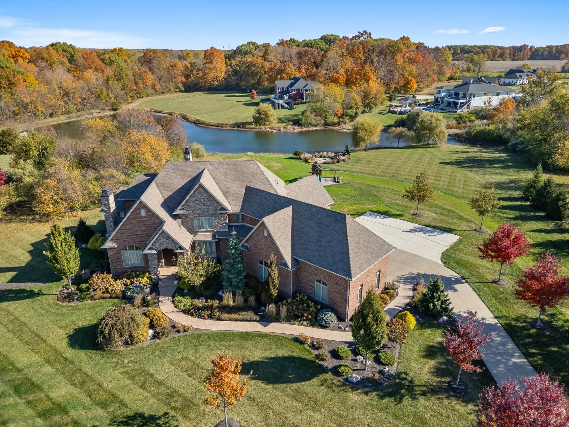 Aerial view of a brick house with a landscaped yard, pond, and autumn trees in a suburban neighborhood