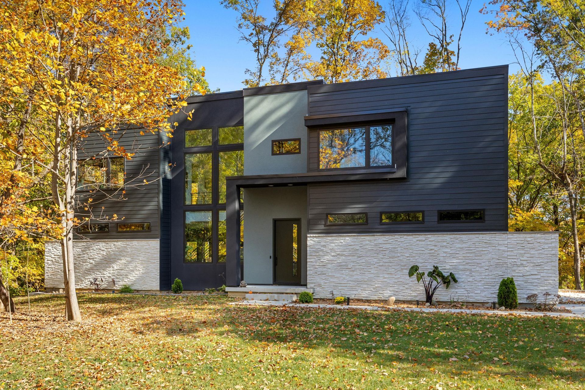 Modern two-story house with dark gray siding, white brick, and autumn trees in front yard