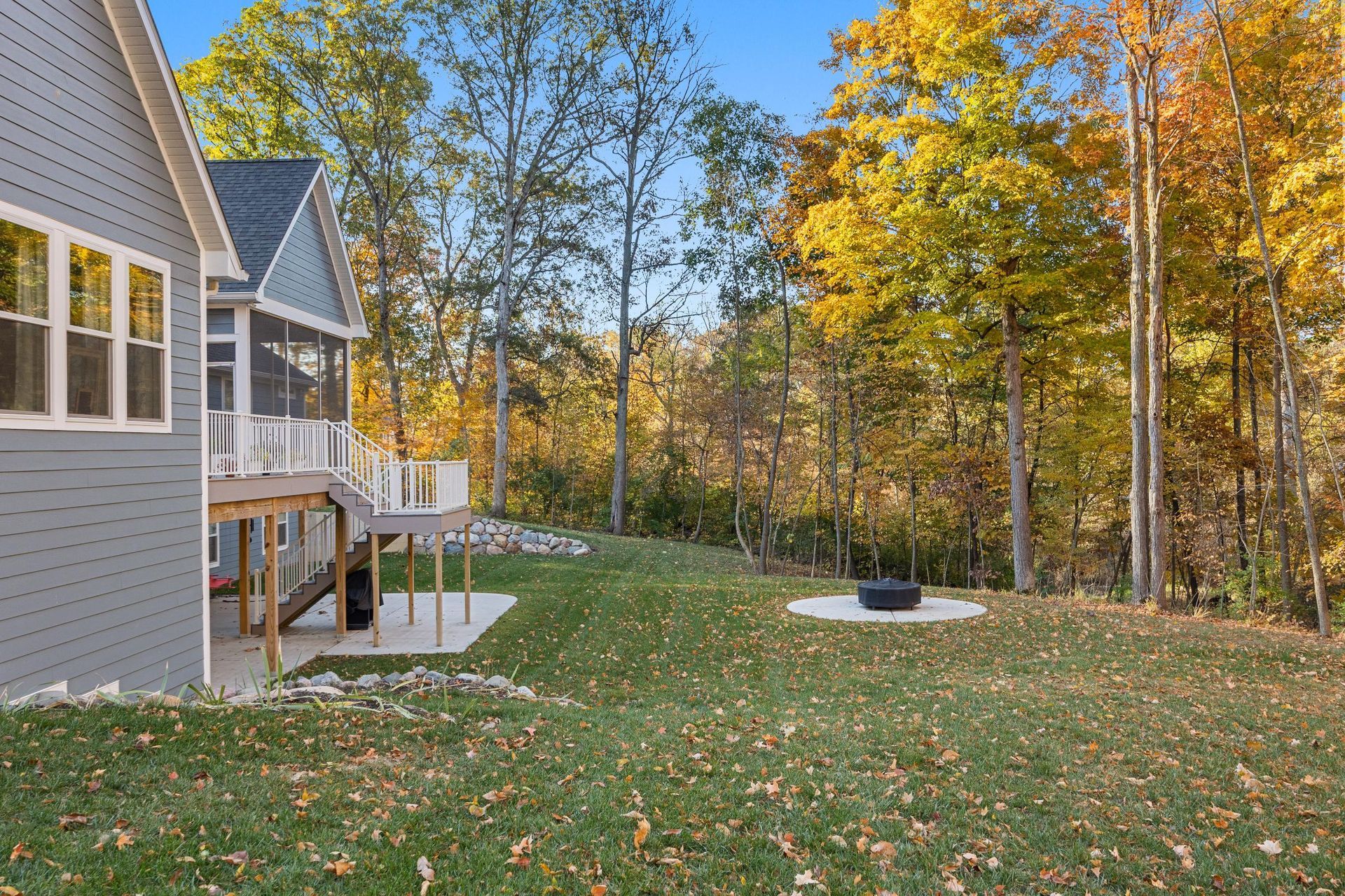 Backyard with gray house, wooden deck, green lawn, and autumn trees around a fire pit