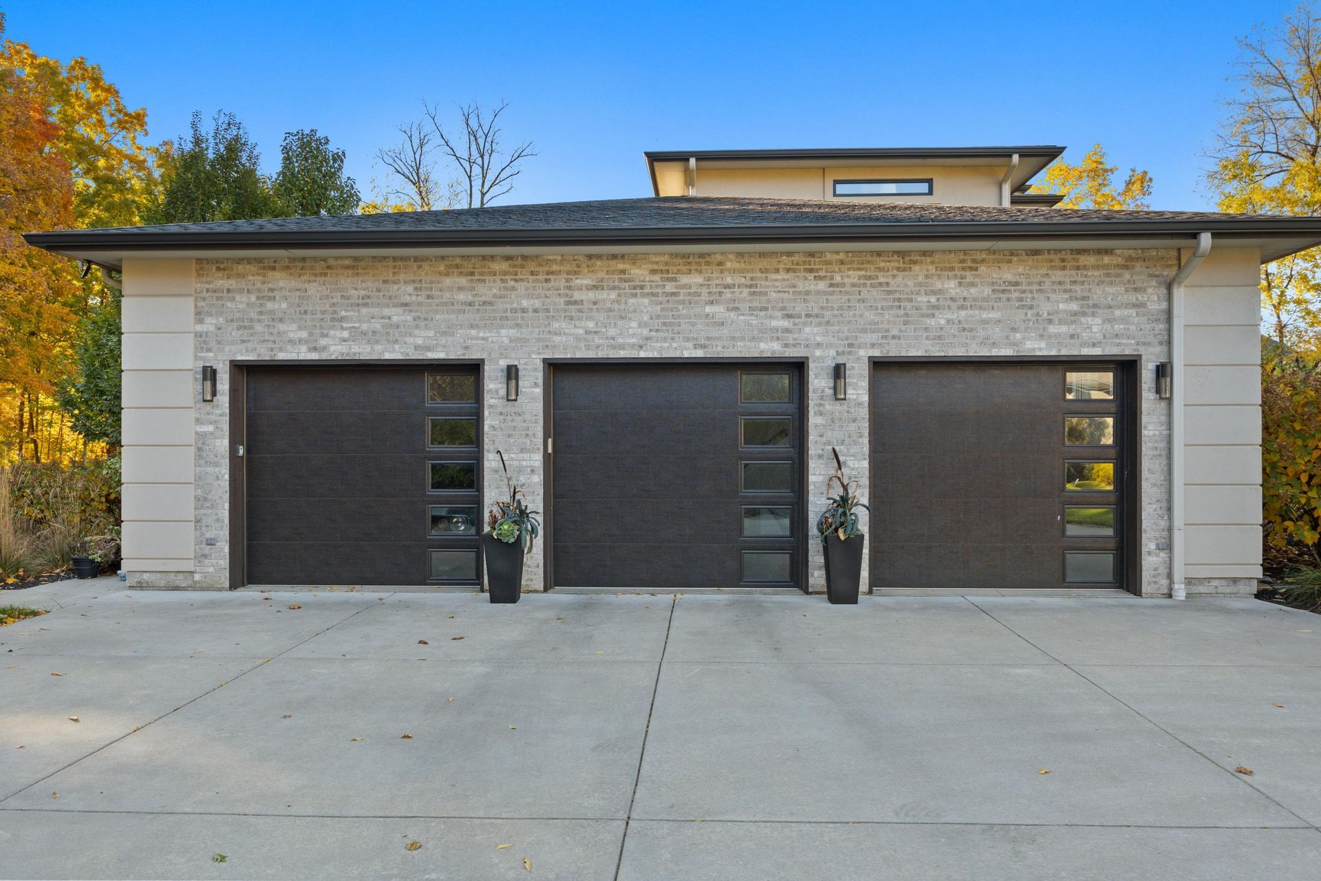 Three black garage doors on a light stone building with a concrete driveway and autumn trees in back