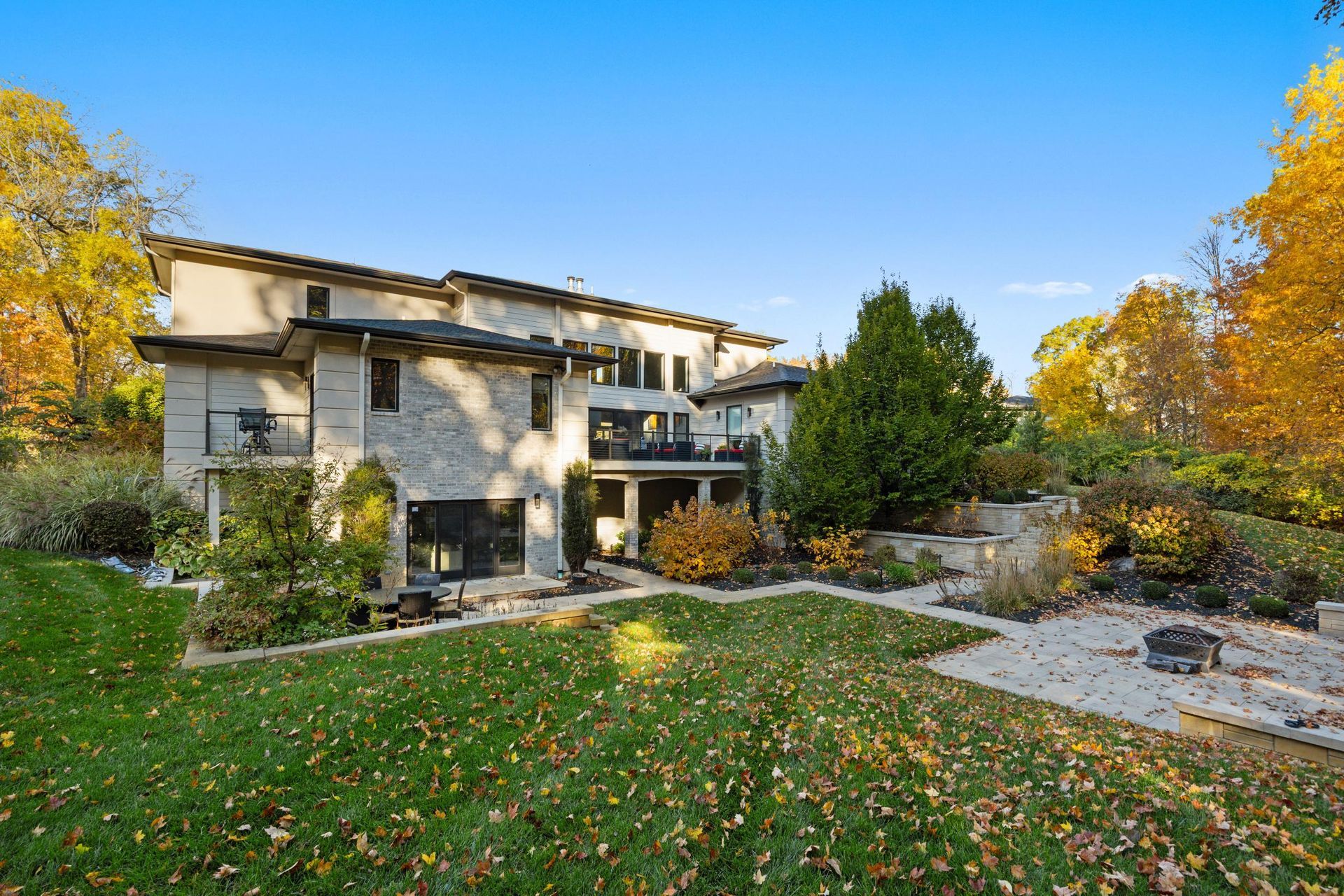 Two-story stone house with autumn trees and a landscaped lawn under a clear blue sky