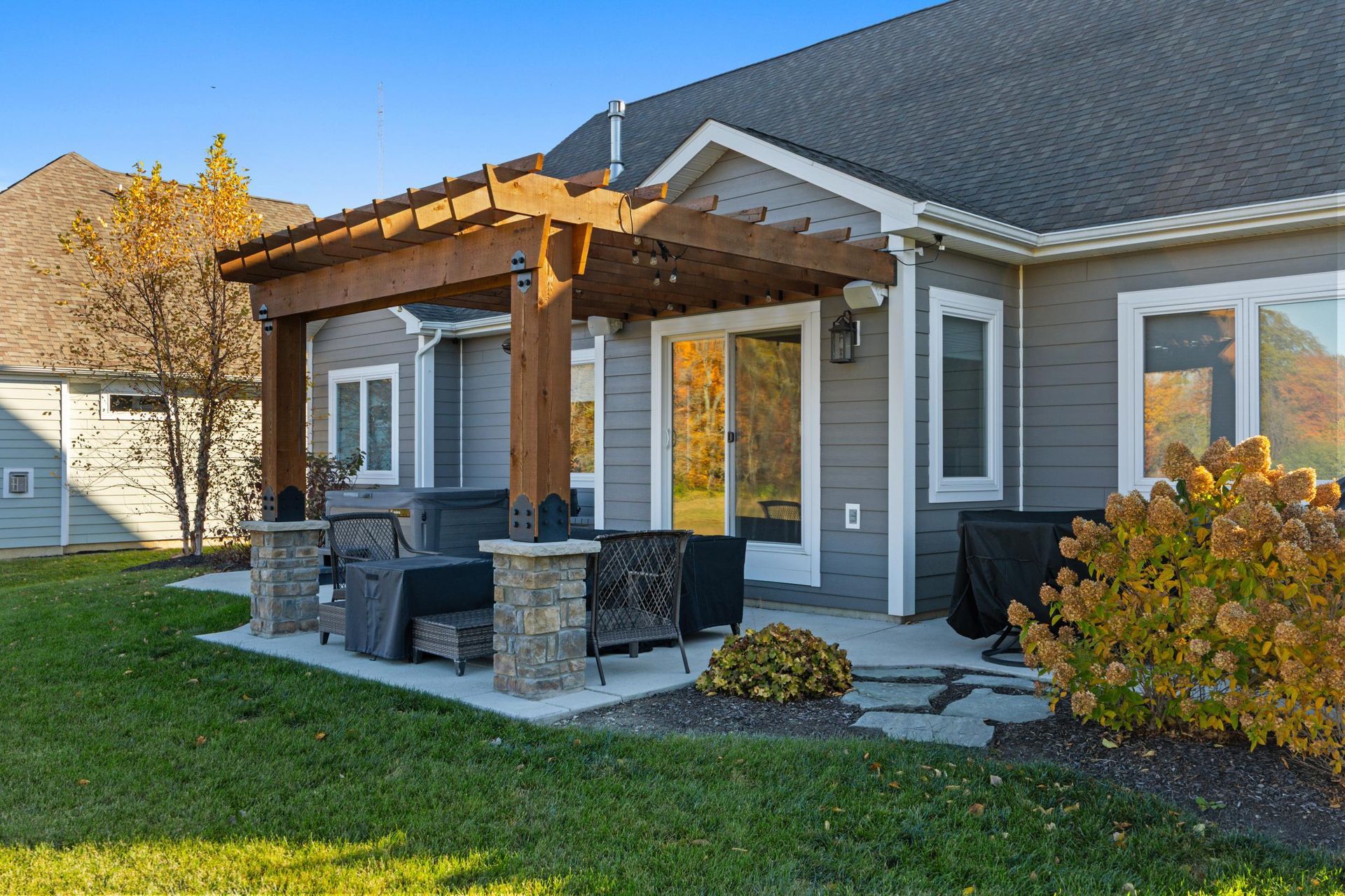 Covered backyard patio with pergola, outdoor dining set, and stone fire pit beside a gray house.