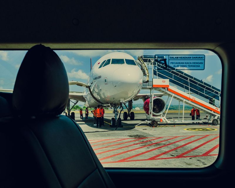 Airplane at airport seen through a car window, with a boarding stairway and ground crew nearby.