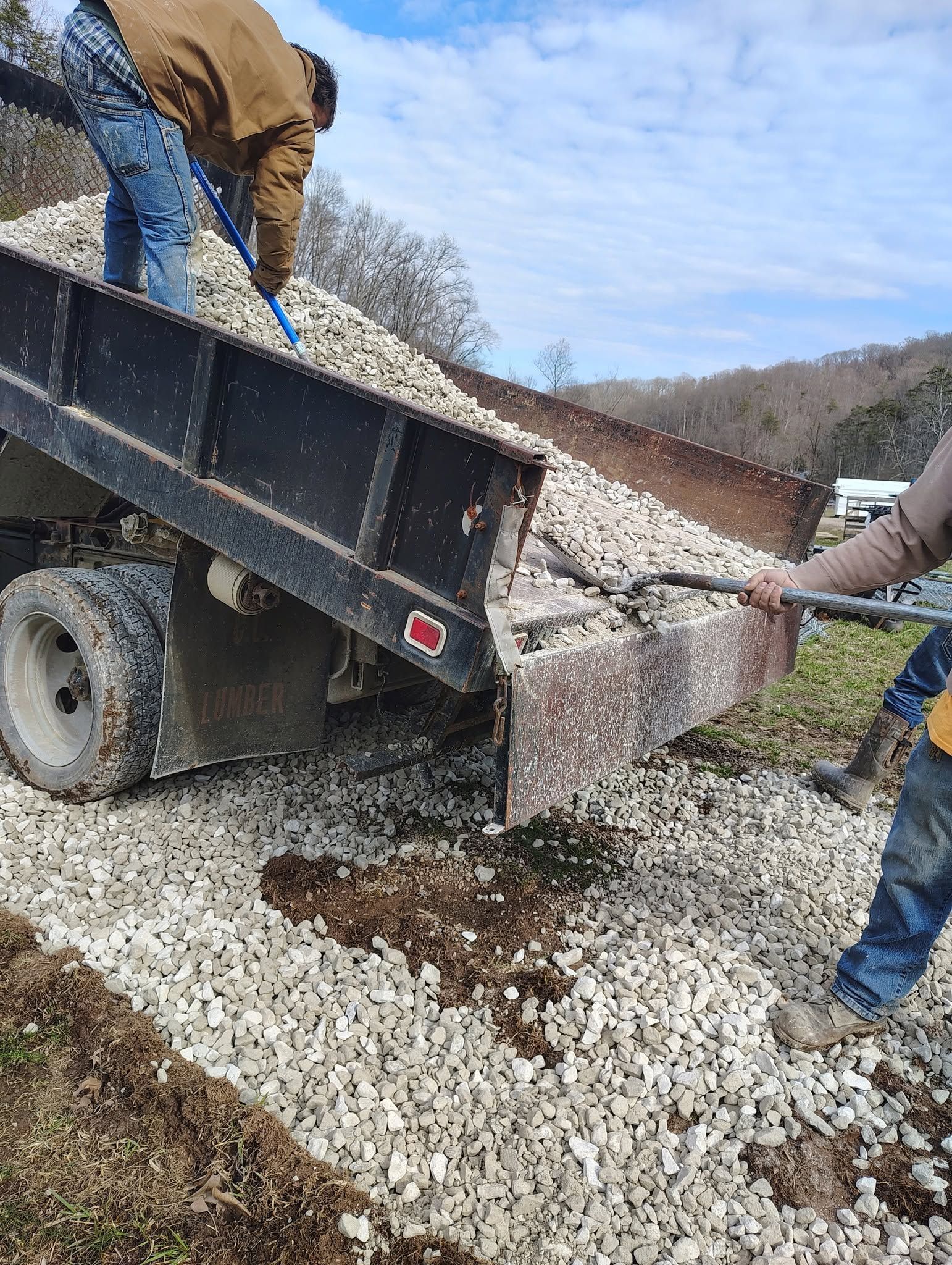 Workers pouring gravel from a truck onto a dirt road in a rural hillside area