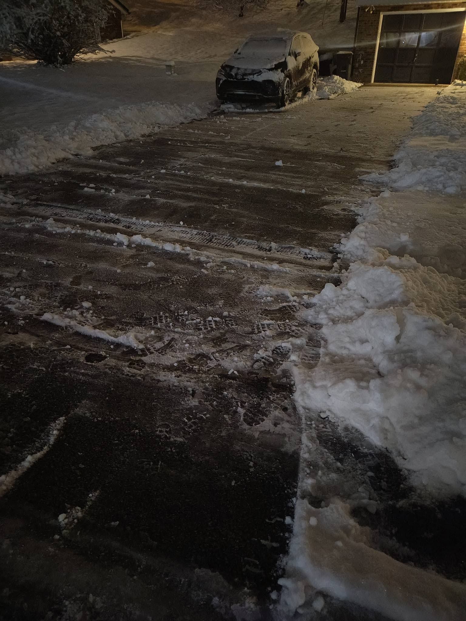 Snow-covered driveway at night with a parked car and garage, lit by porch lights.