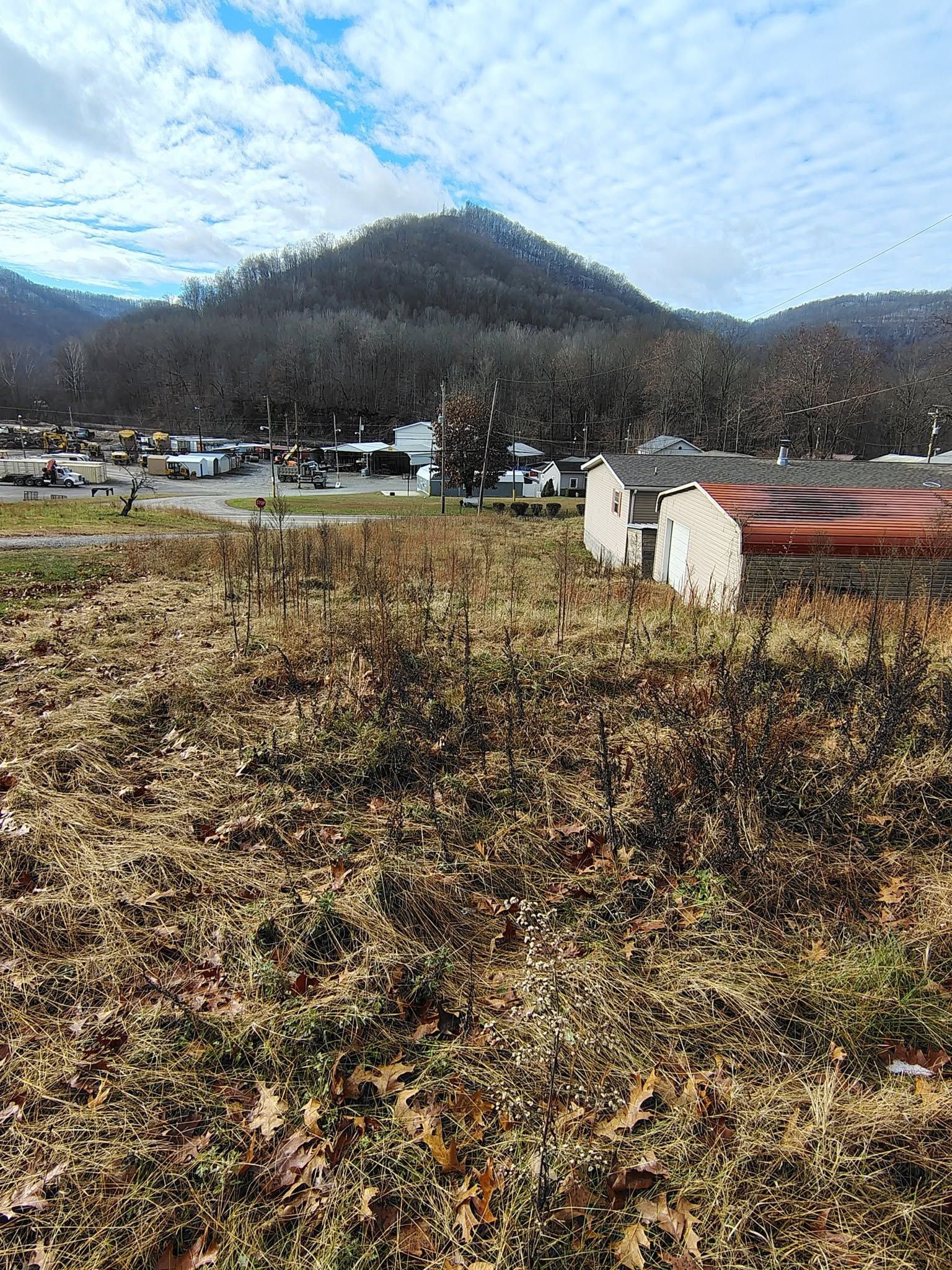 Grassy field with white barriers, road, and a wooded hill under a cloudy sky