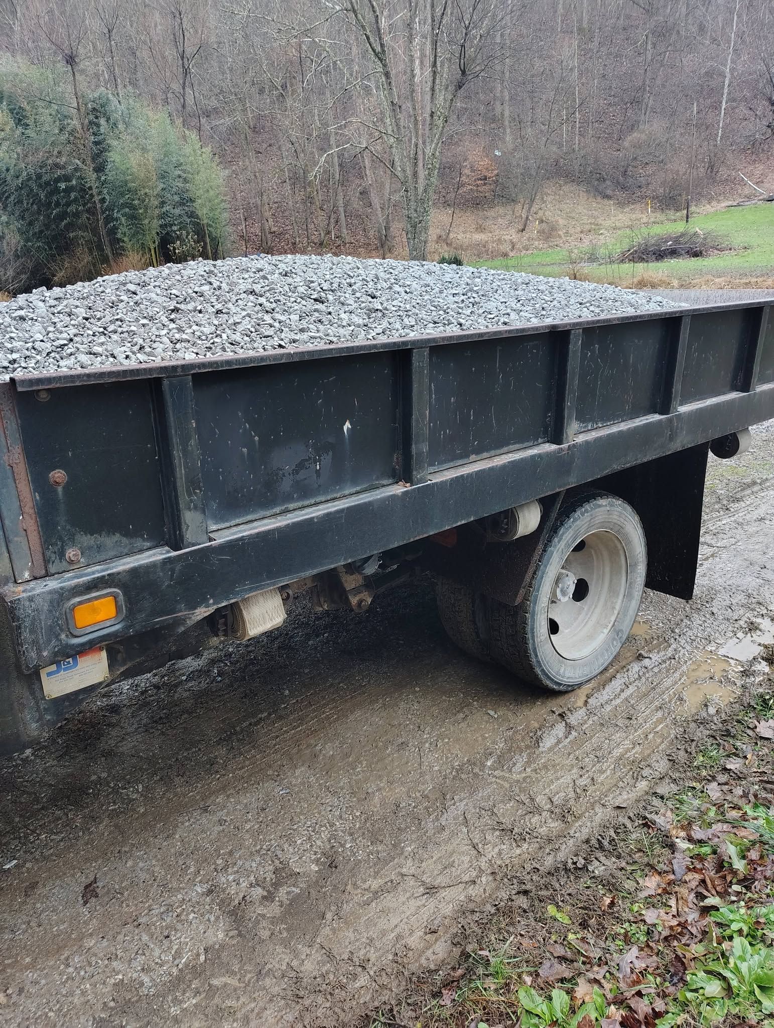 Black utility trailer parked on a gravel path in a wooded area, loaded with rocks.