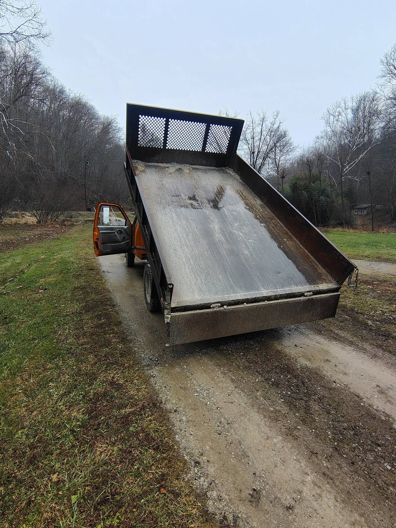 Orange dump truck tipping a load onto a muddy dirt road in a wooded area