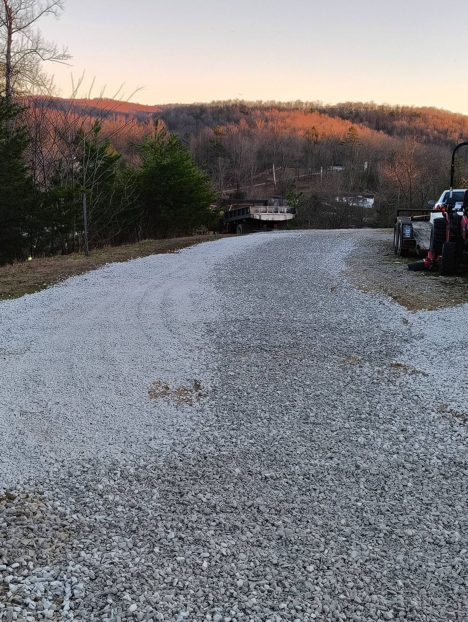 Gravel road leading toward wooded hills at sunset, with a small vehicle parked on the right.