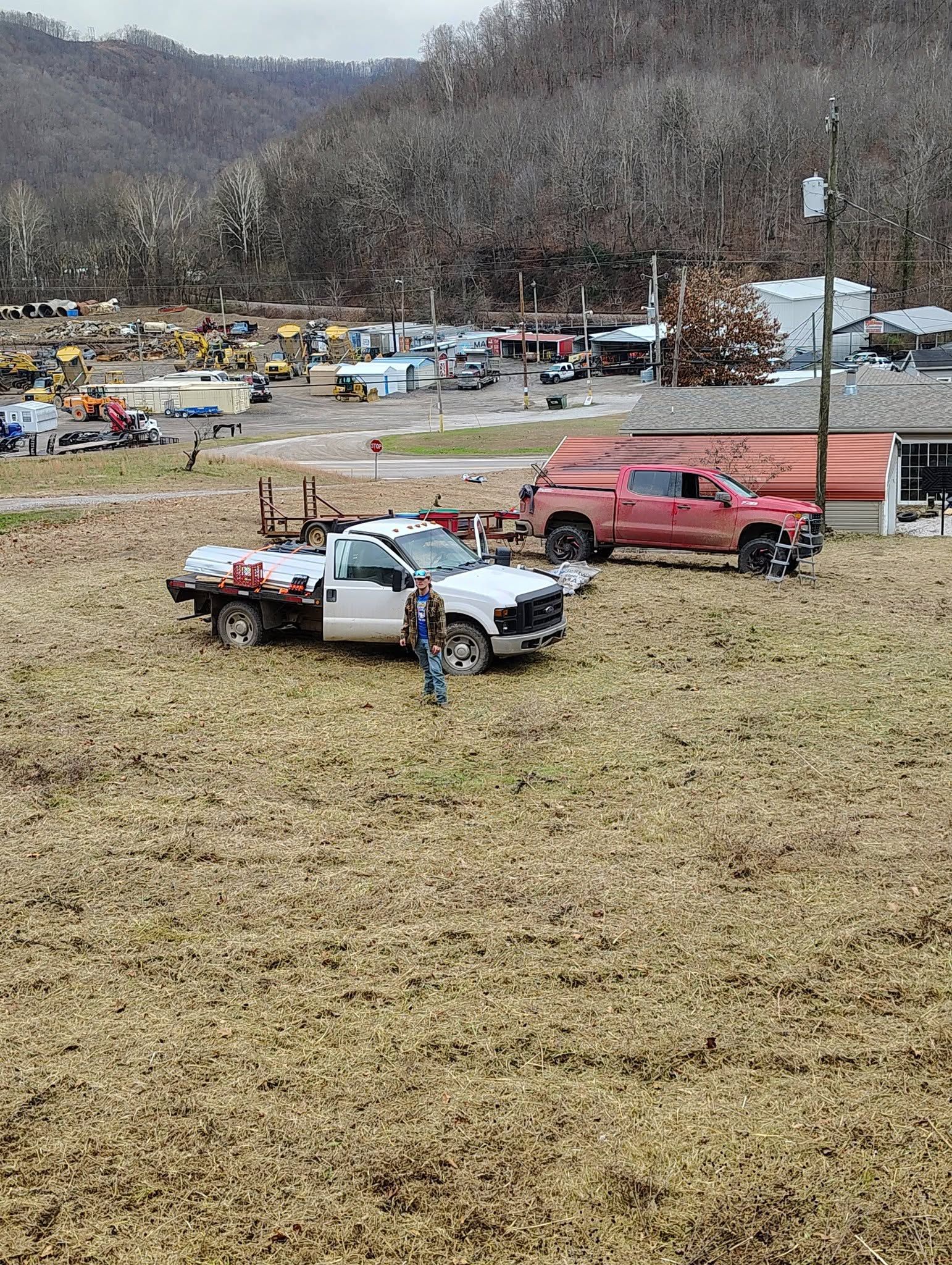 People at an outdoor muddy truck gathering with several parked pickup trucks and a wooded hillside backdrop