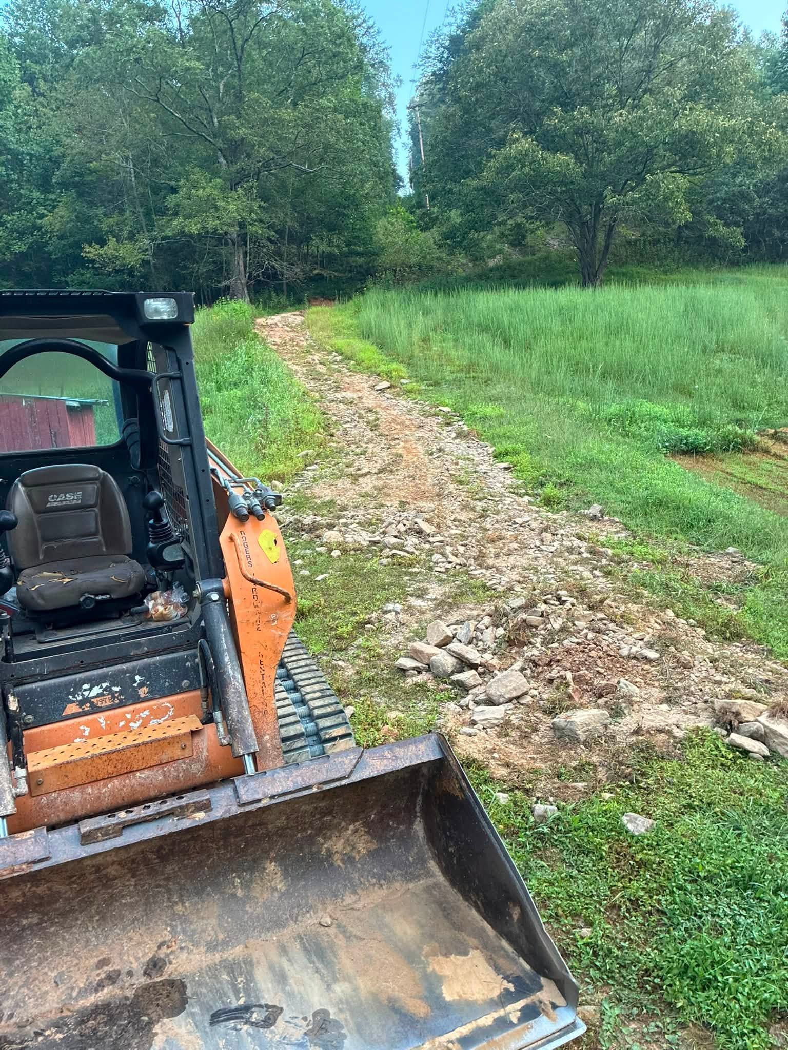 Orange skid steer beside a rocky path in a grassy field with trees in the background