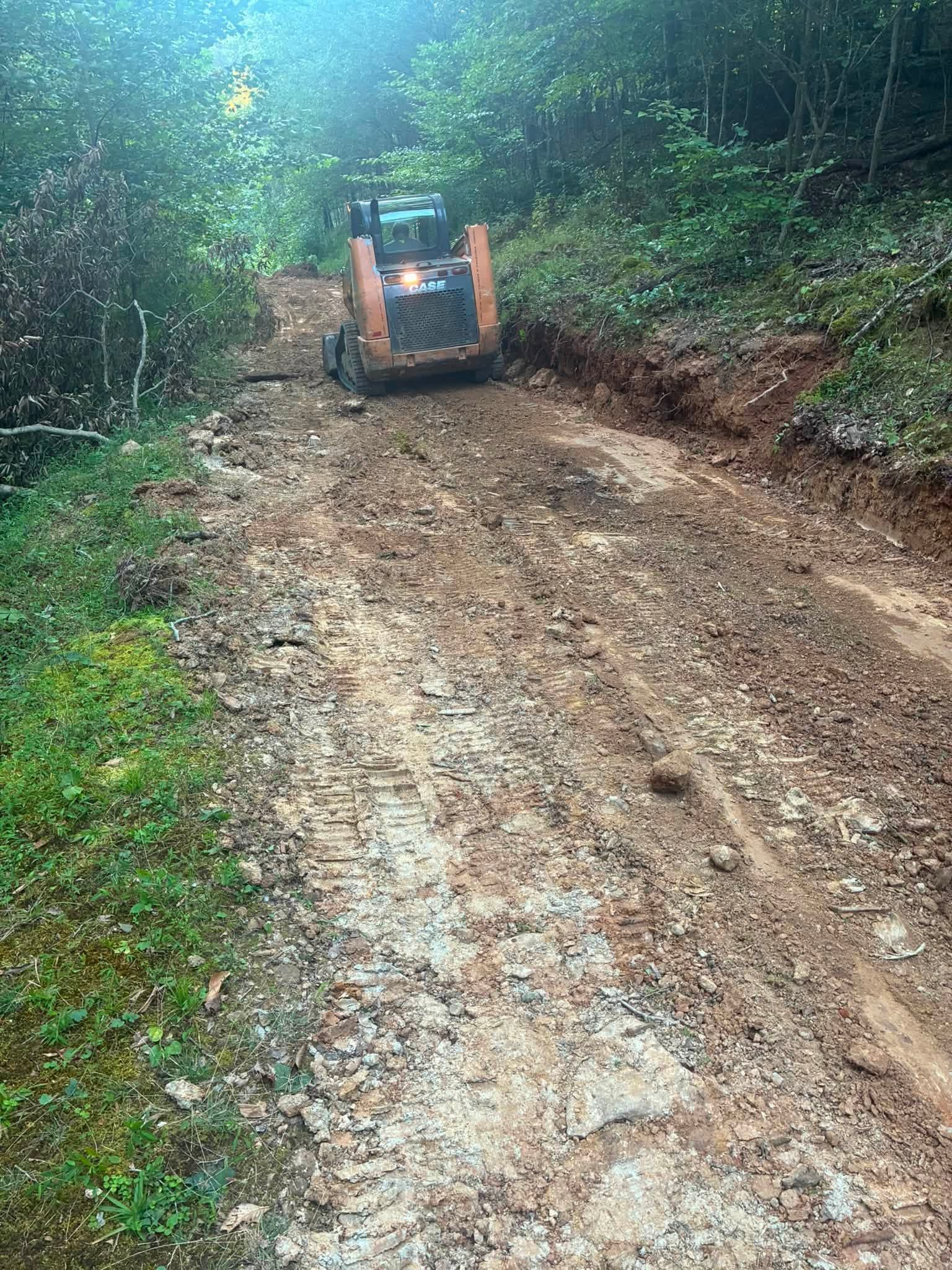 Orange skid steer on a muddy forest road, grading a dirt path through dense green woods.