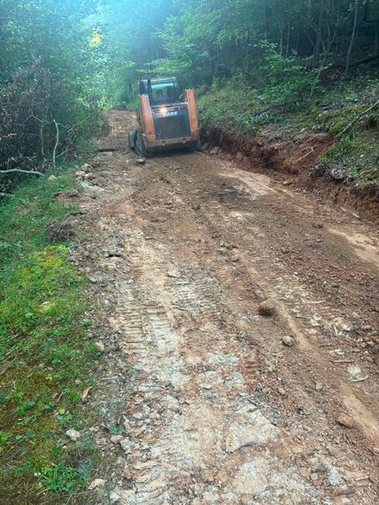Orange skid steer on a muddy forest road, grading a dirt path through dense green woods.