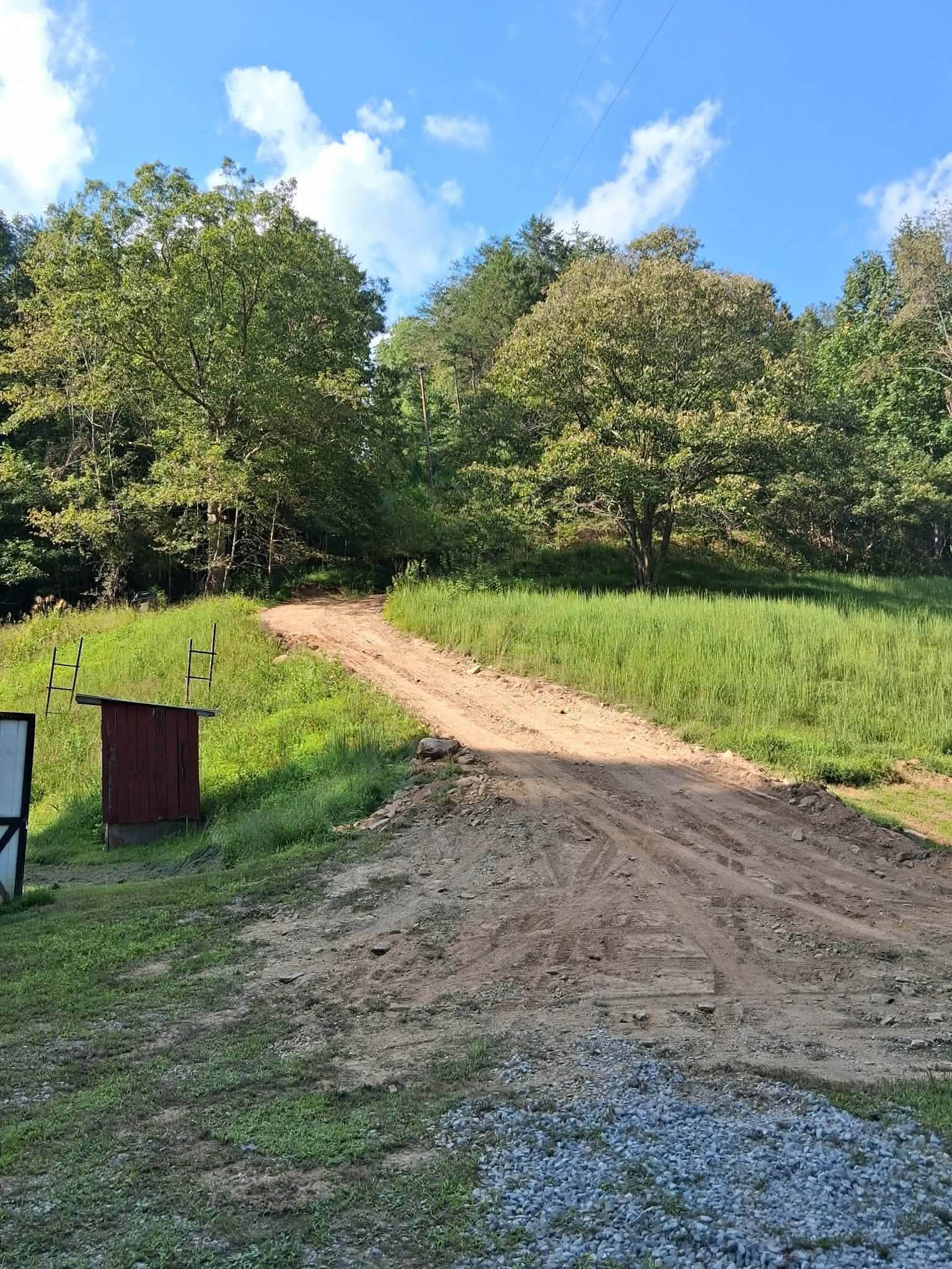 Dirt path through a grassy field toward trees under a blue sky with clouds, with a trash bin at left.