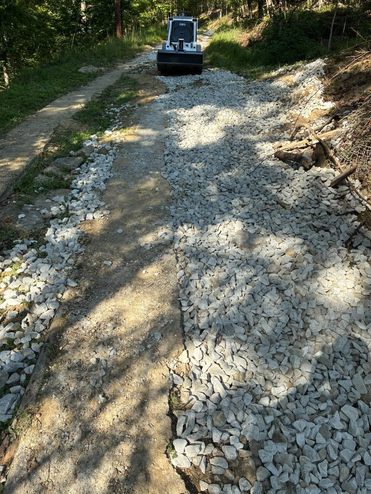 Gravel road under repair with a small excavator ahead, surrounded by trees and dappled sunlight