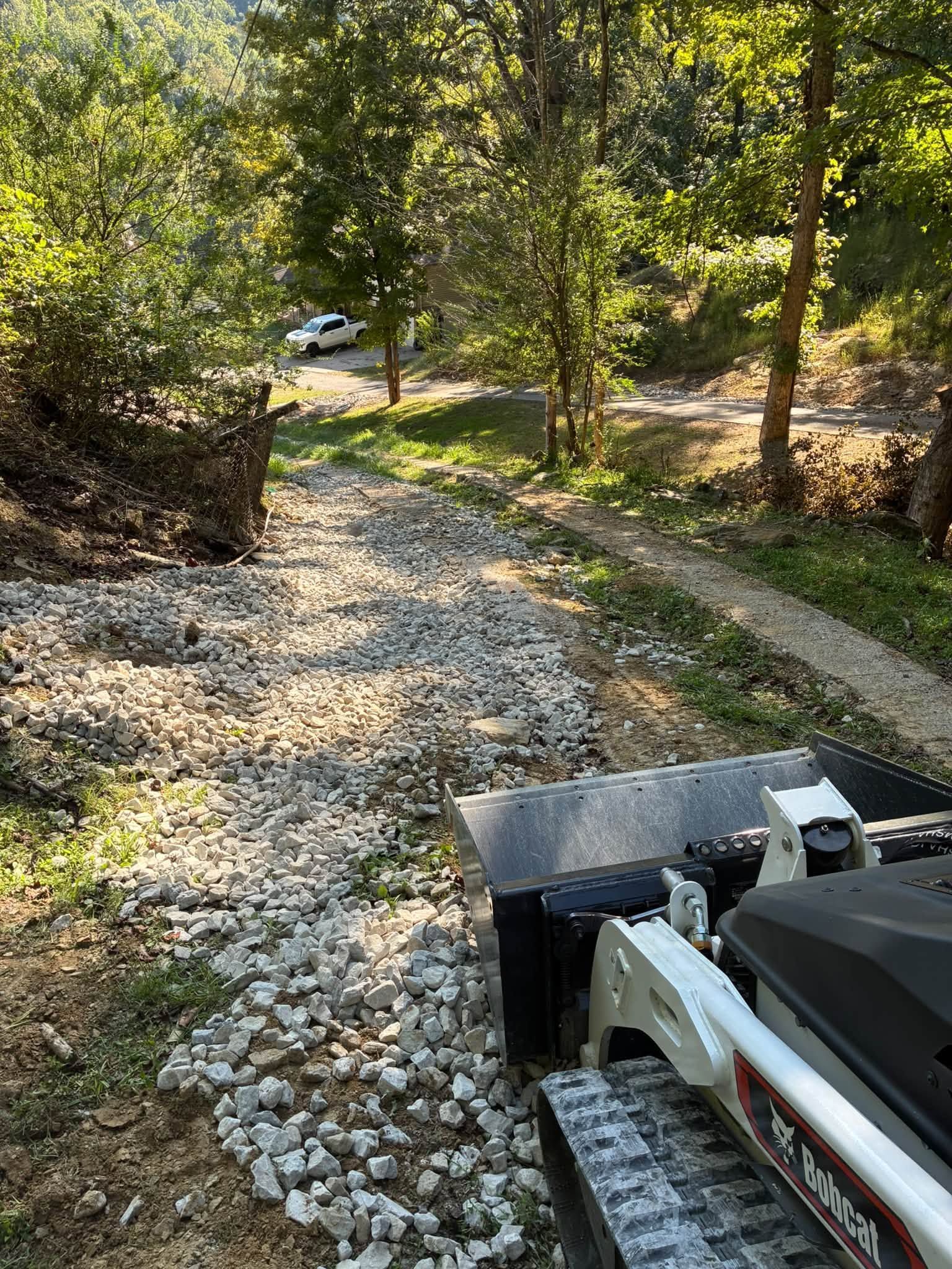 Skid-steer clearing a rocky gravel path through a wooded area in bright sunlight