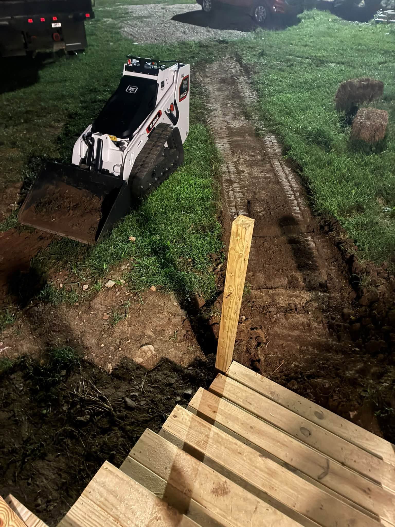 Small white skid-steer on muddy grass beside a dirt track and wooden steps at night