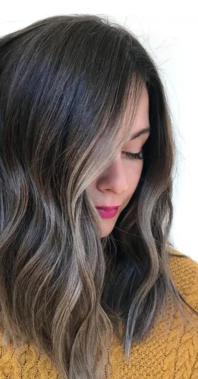 Primo piano di una persona con lunghi capelli scuri e mossi e rossetto rosa, che guarda in basso indossando un maglione color senape.