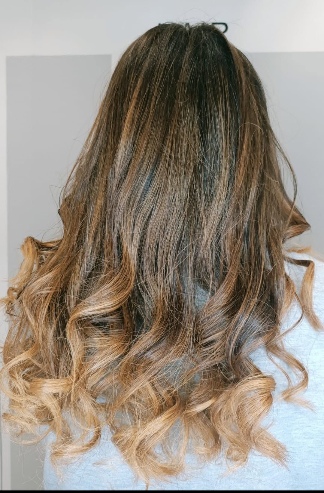 Vista posteriore di capelli mossi castano scuro con punte bionde sfumate su sfondo grigio chiaro.