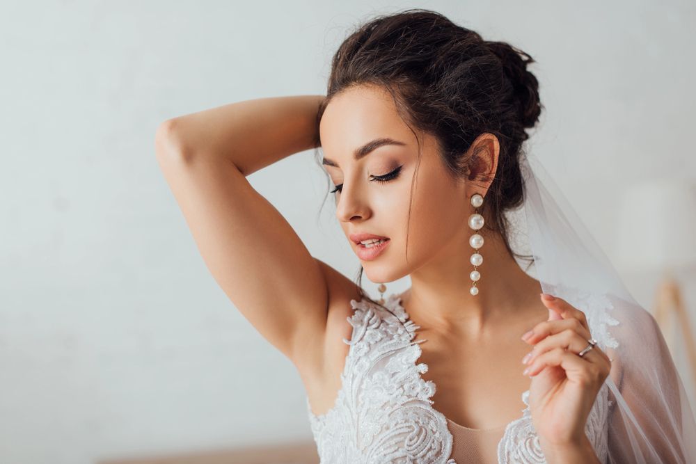 Sposa in abito di pizzo bianco che si sistema un orecchino di perle pendente, con un velo trasparente e uno sfondo neutro e delicato.