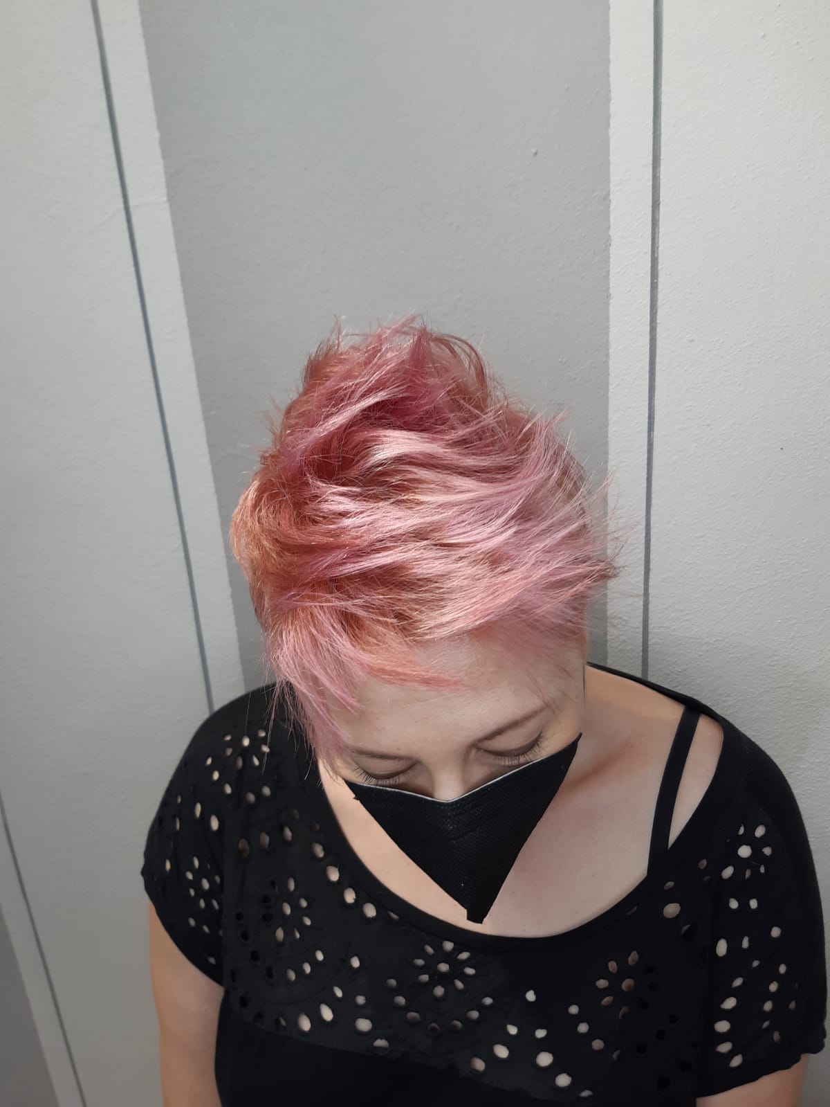Persona con i capelli corti rosa, che guarda in basso, con indosso una maglietta nera, davanti a una parete chiara.