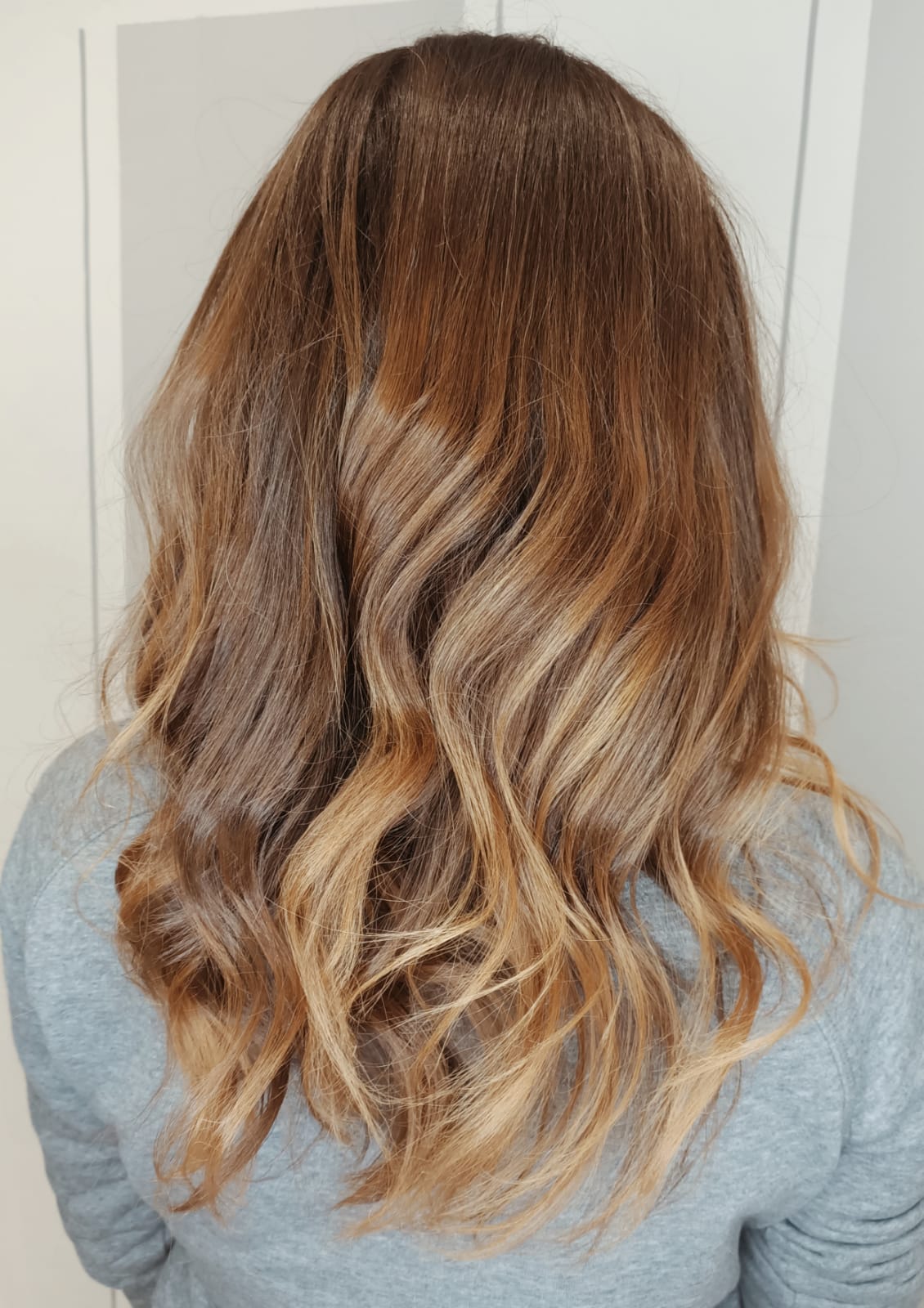 Vista posteriore di lunghi capelli castani ondulati con punte bionde sfumate, sopra una camicia grigia.