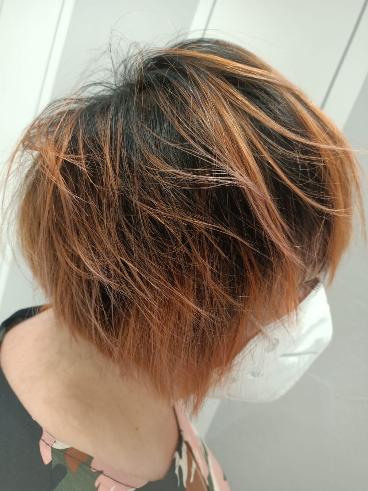 Vista laterale di una persona con capelli corti a strati castani e arancioni che indossa una mascherina bianca.