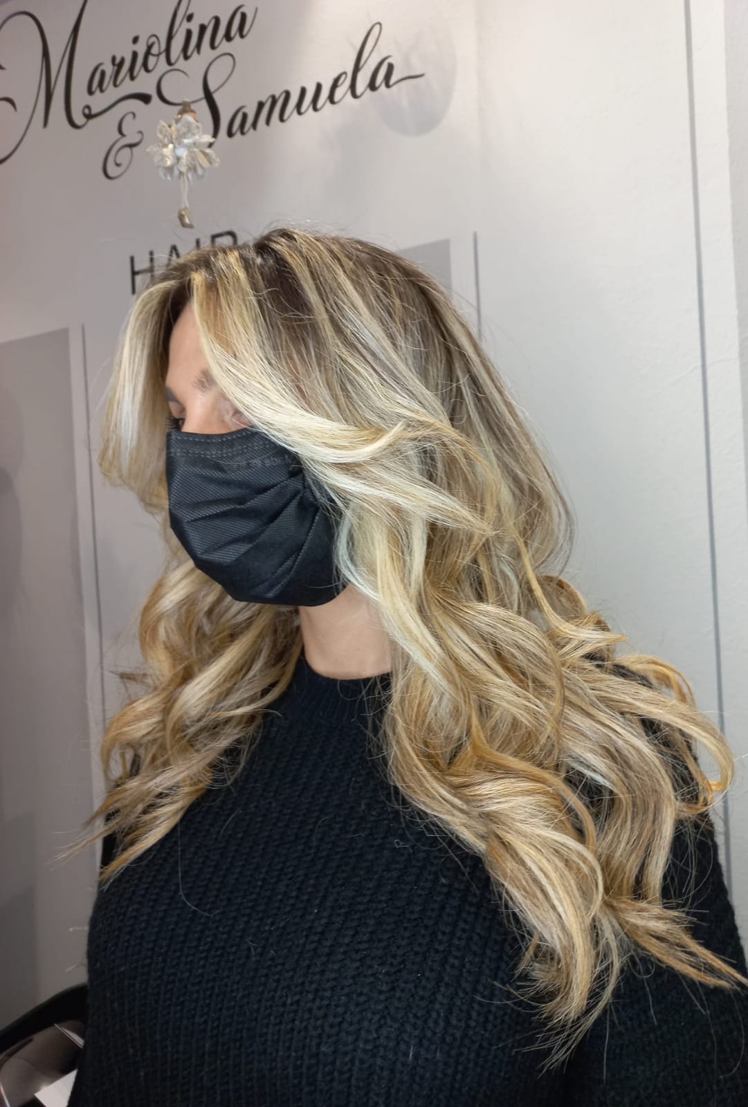 Donna con lunghi capelli biondi e mascherina nera in un salone, indossa un maglione nero.