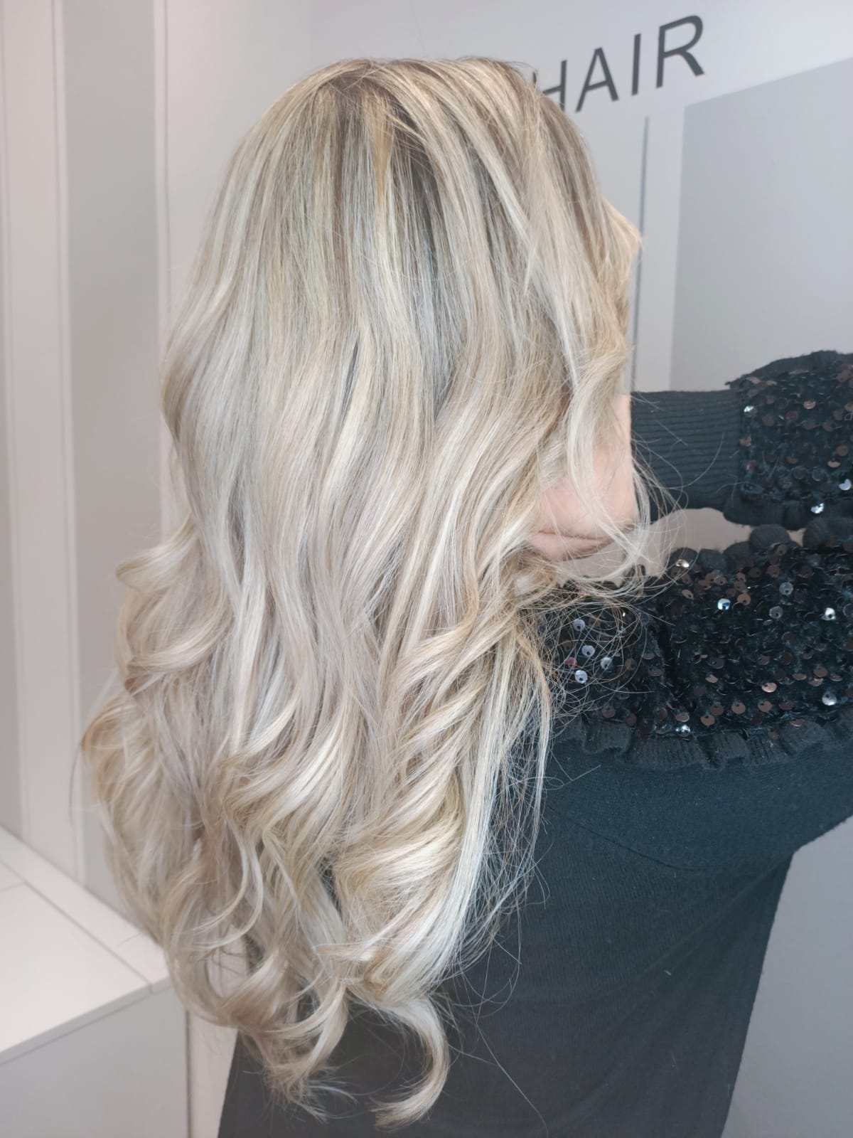 Vista posteriore di lunghi capelli biondi ondulati con radici più scure e riflessi argentati in un salone di bellezza.