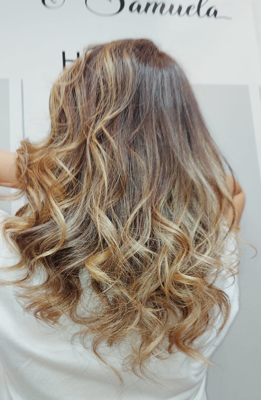 Capelli lunghi, mossi e biondi con balayage, visti da dietro su uno sfondo chiaro.