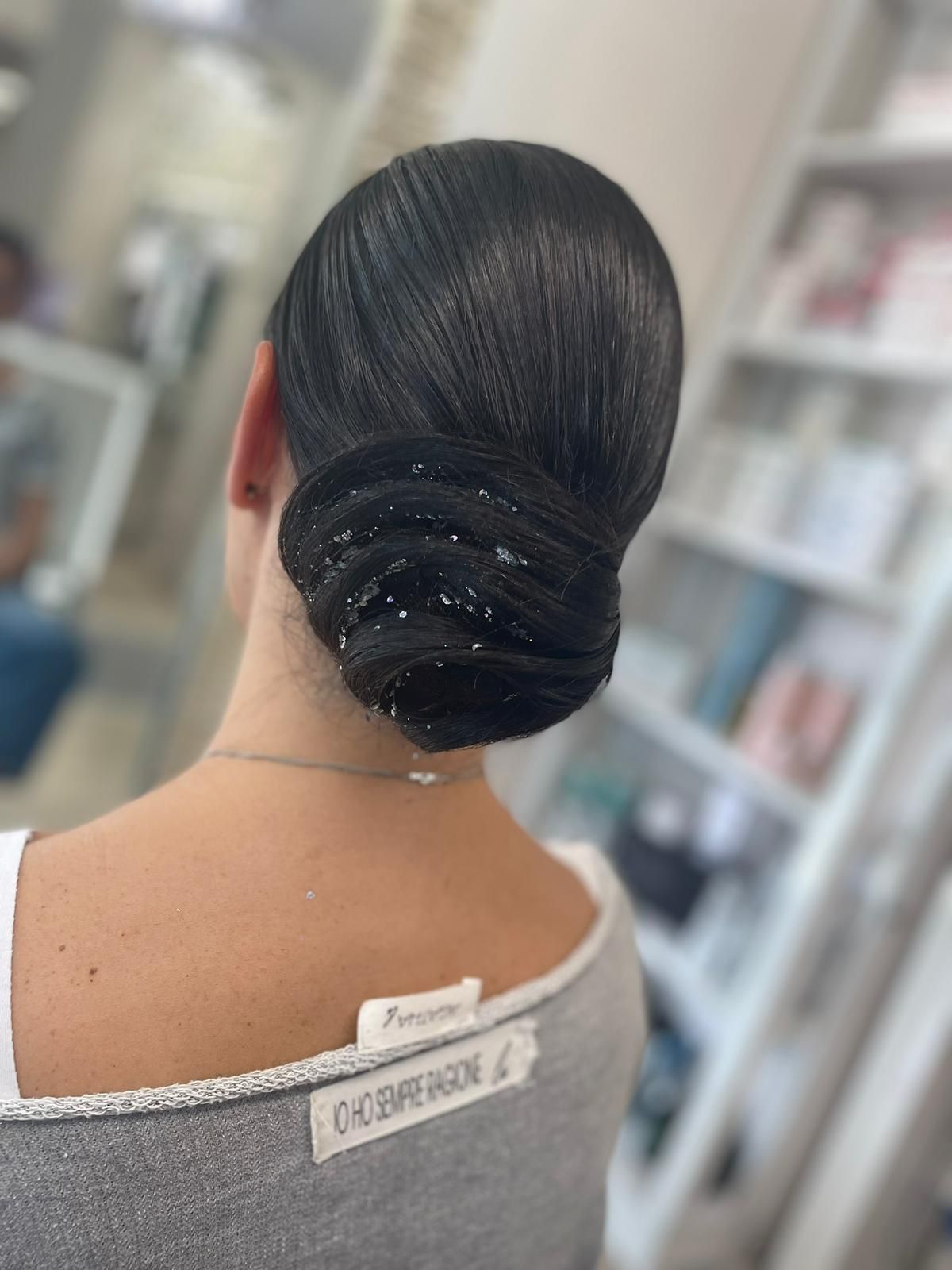 Vista posteriore di una persona con uno chignon basso ed elegante seduta su una poltrona da parrucchiere, che indossa un top grigio a spalle scoperte.