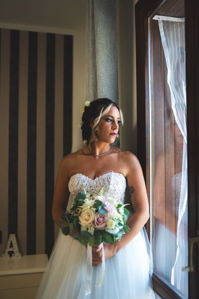 La sposa, in abito bianco, tiene in mano un bouquet dai colori pastello, in piedi accanto a una finestra con tende trasparenti.