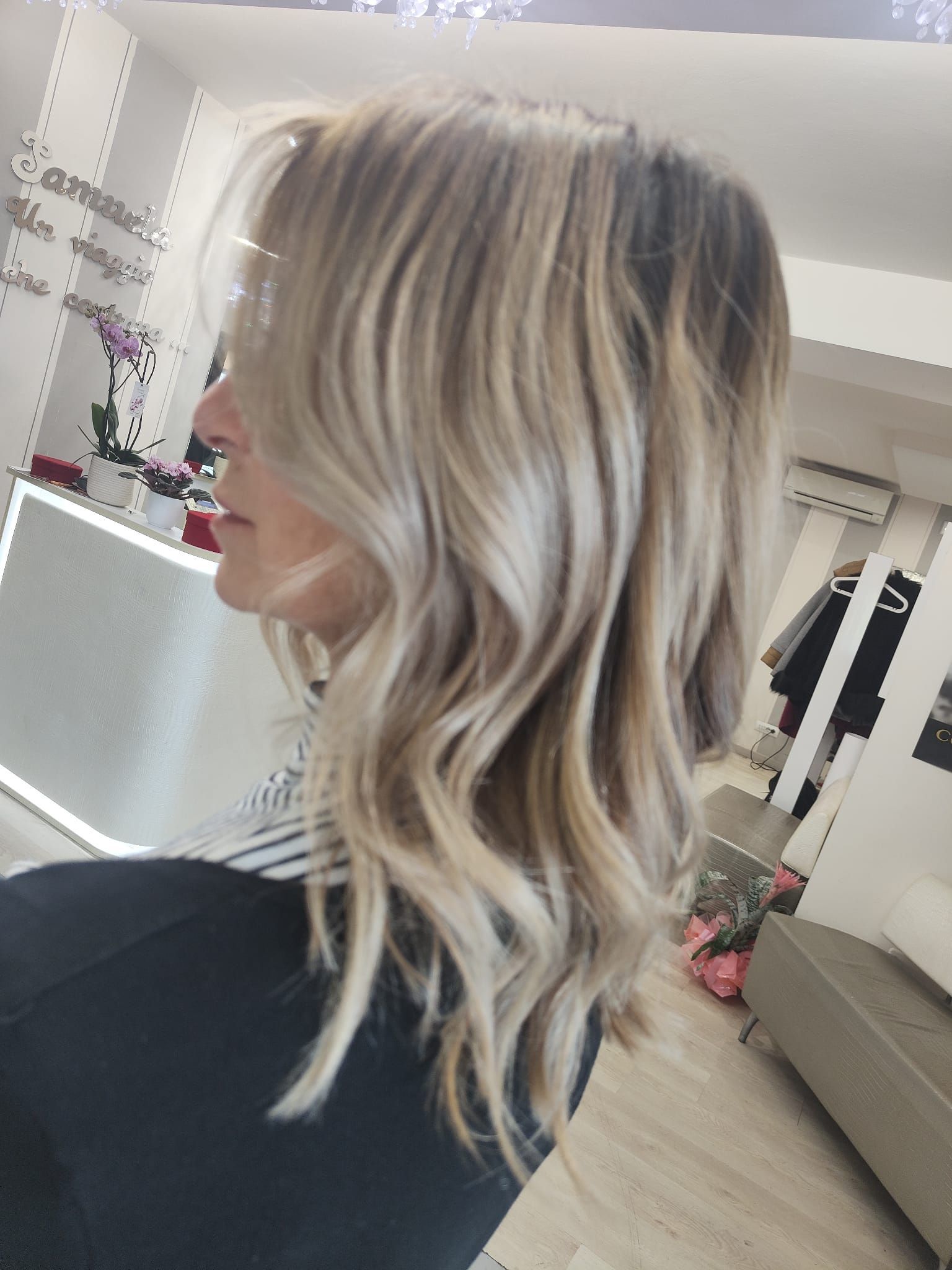 Profilo laterale di una donna con capelli biondi ondulati con mèches, in un luminoso salone di bellezza al chiuso.