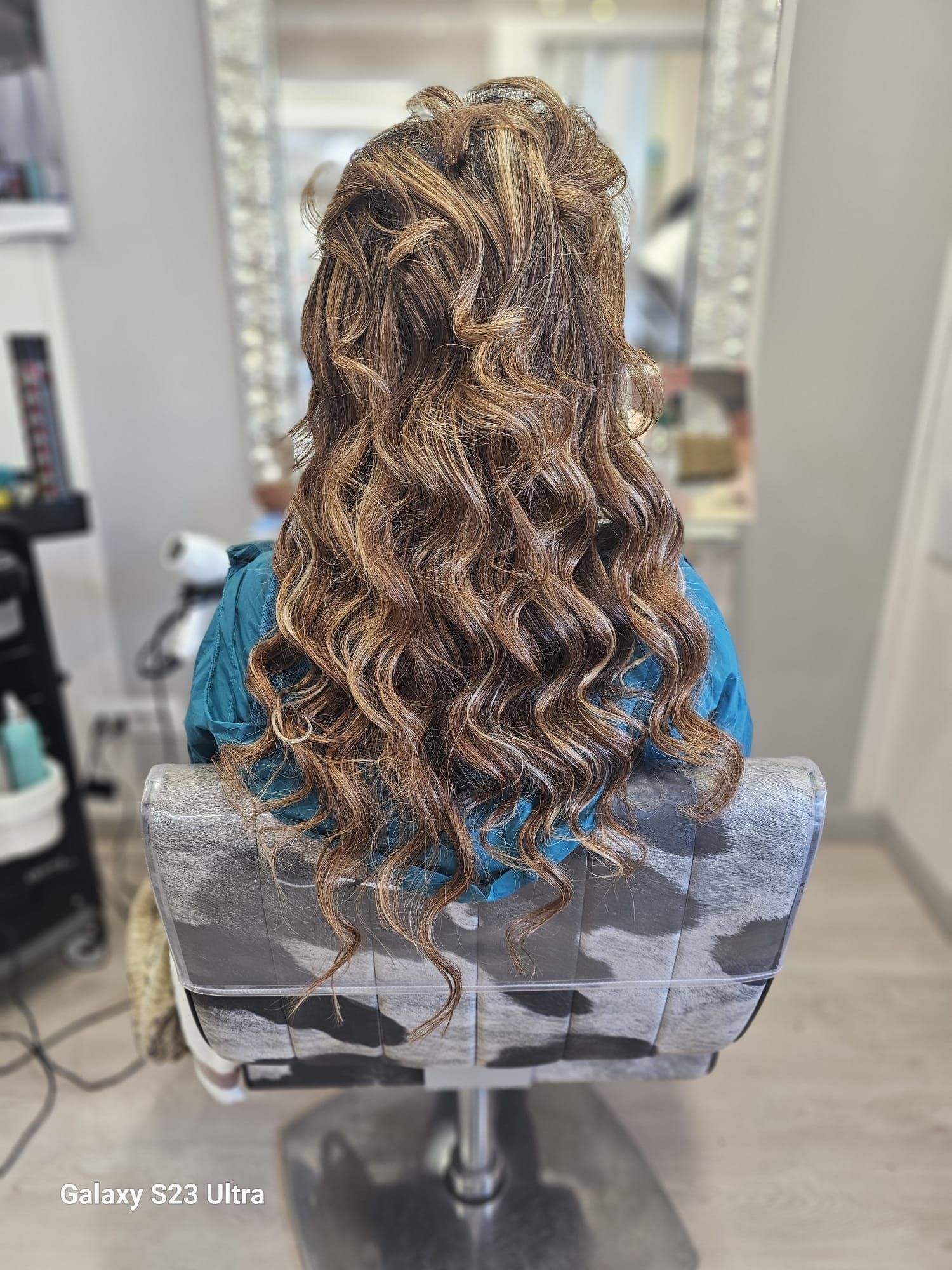 Vista posteriore di una persona con lunghi capelli ricci con mèches, seduta su una poltrona da parrucchiere.