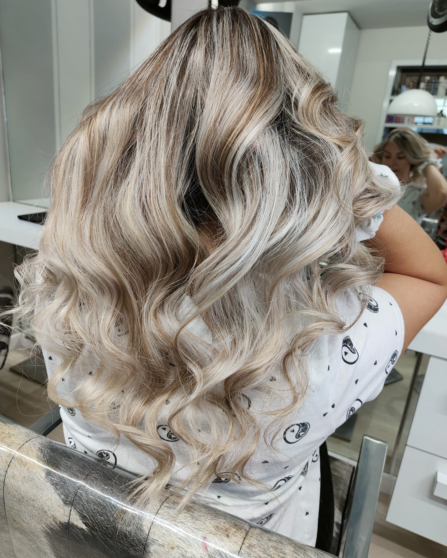 Capelli biondi mossi con balayage visti da dietro, seduti su una poltrona da salone, con specchio e prodotti a portata di mano.