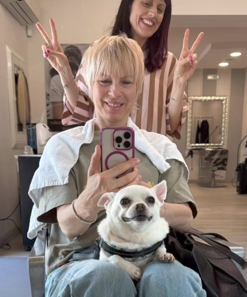 Selfie allo specchio del salone con una donna sorridente, un'altra persona che fa il segno della pace e un cagnolino con un mantello.