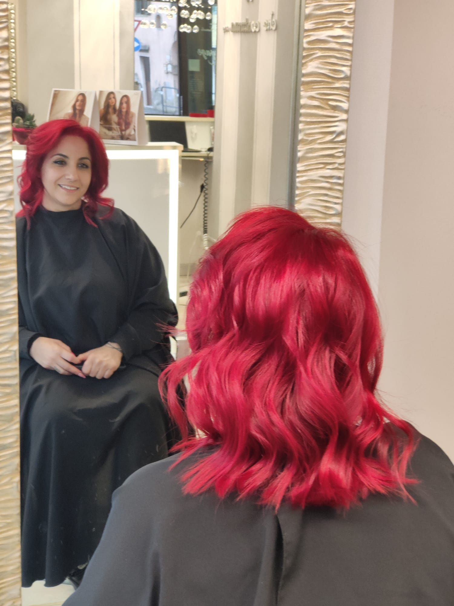 Una donna dai capelli rosso acceso è seduta su una poltrona da parrucchiere, riflessa in uno specchio con indosso un mantello nero.