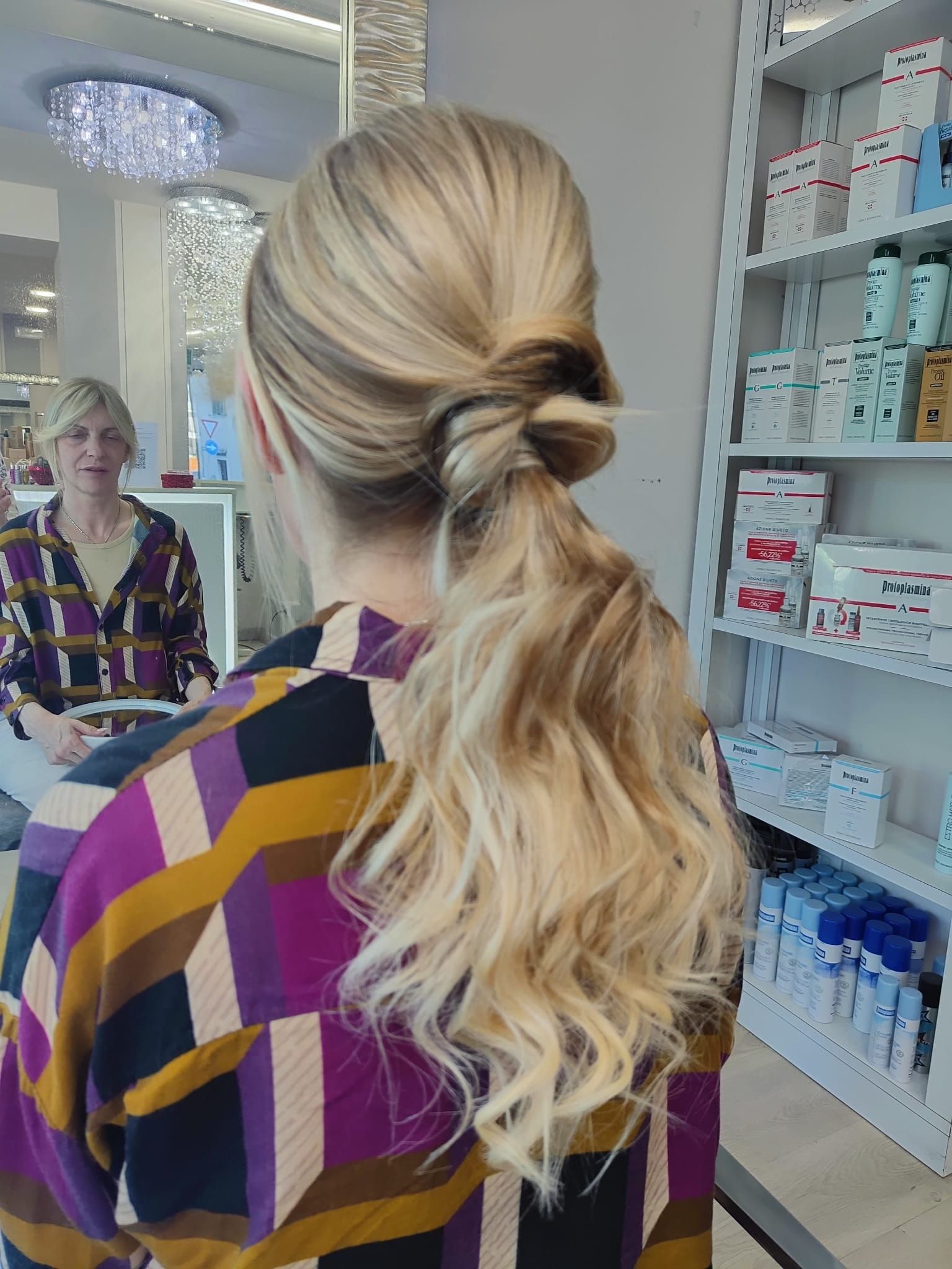 Donna con coda di cavallo bionda in un salone, con indosso una camicetta viola e gialla, vista di spalle davanti a uno specchio.