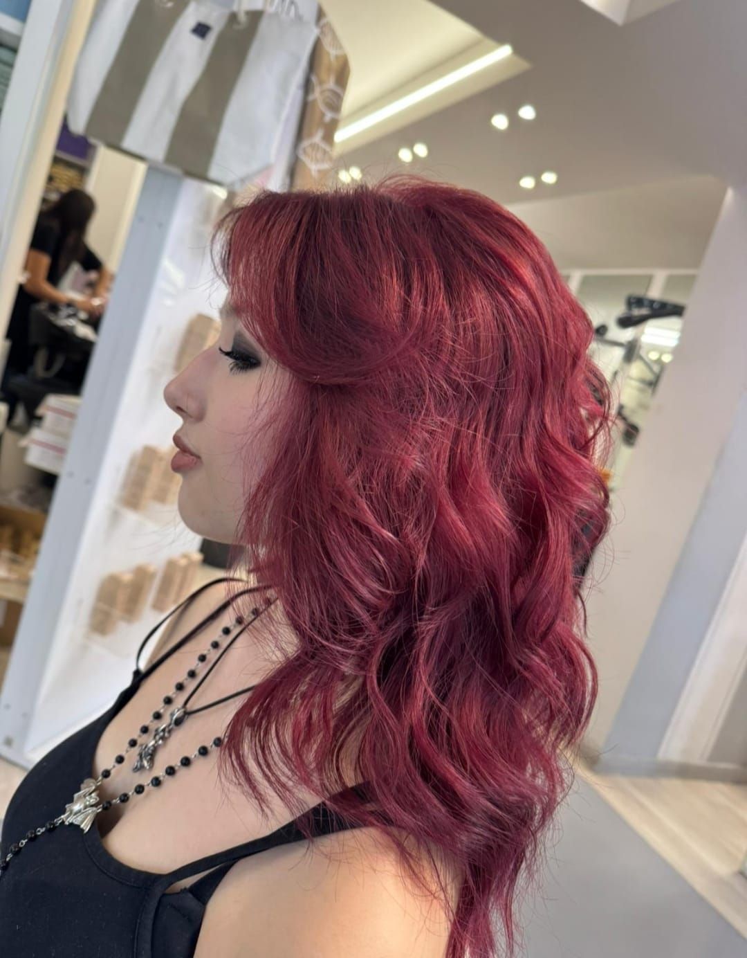 Profilo laterale di una persona con capelli mossi color magenta che indossa una maglietta nera in un negozio di abbigliamento luminoso.
